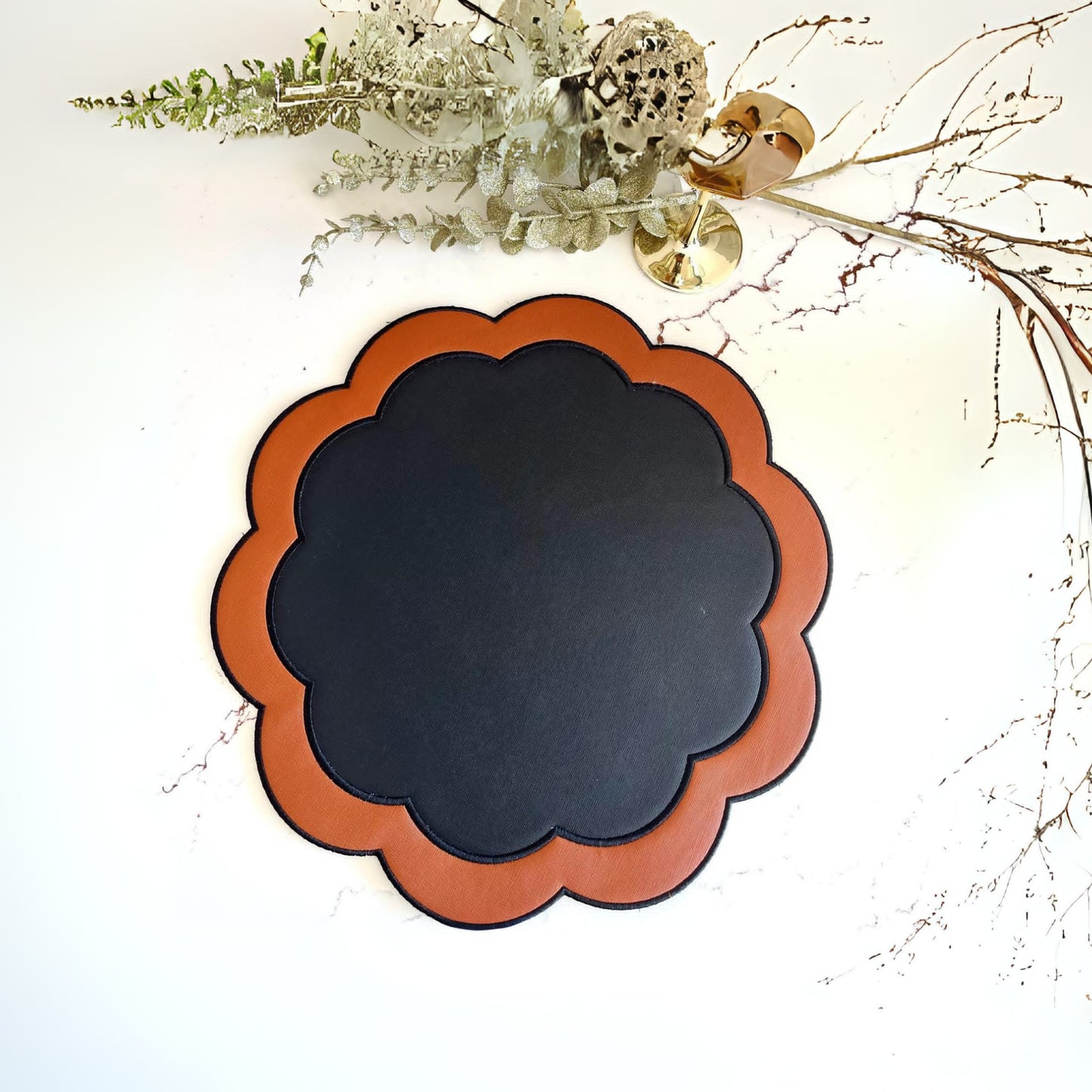 A round black faux leather placemat with an orange border, displayed against a neutral background with some decorative foliage at the top.