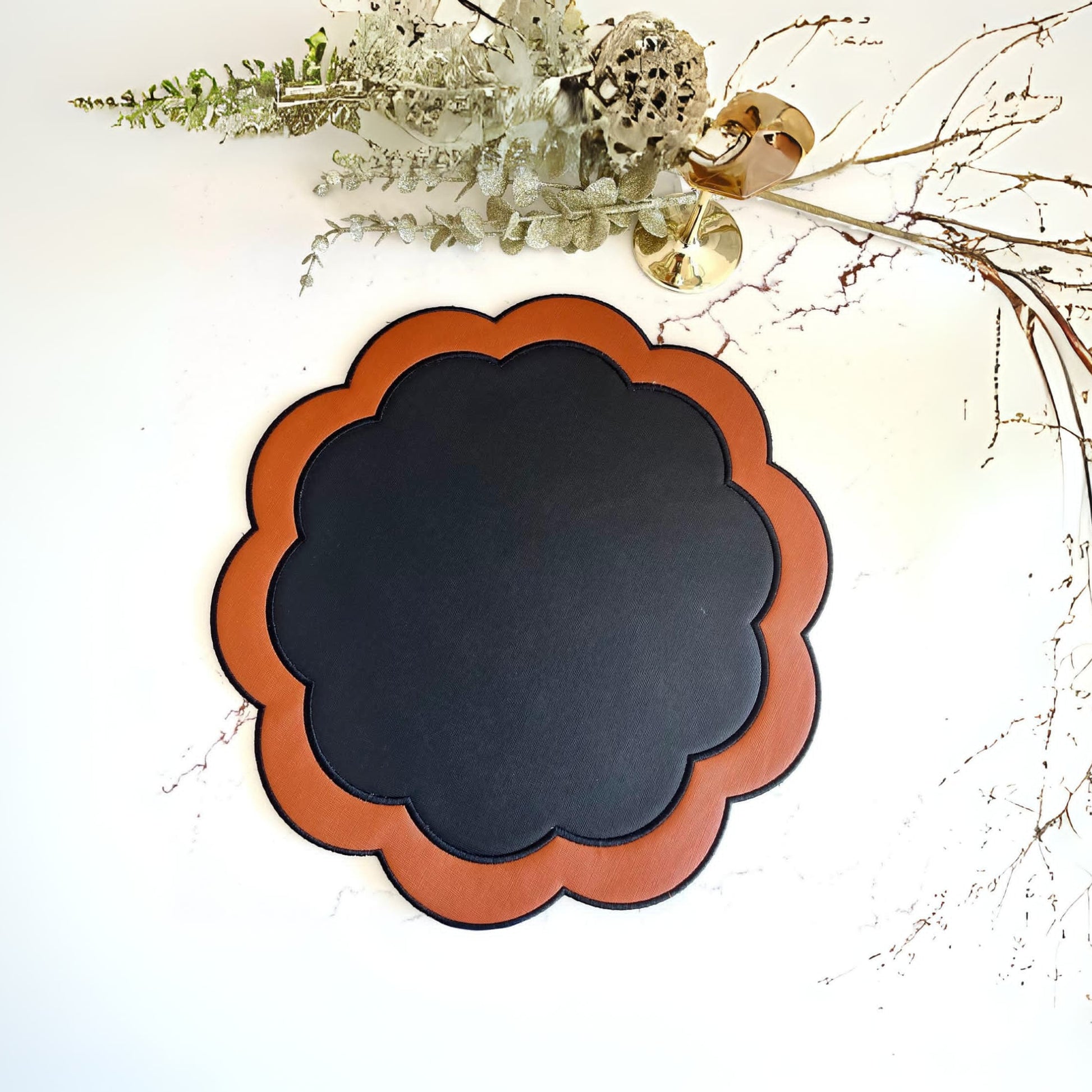 A round black faux leather placemat with an orange border, displayed against a neutral background with some decorative foliage at the top.