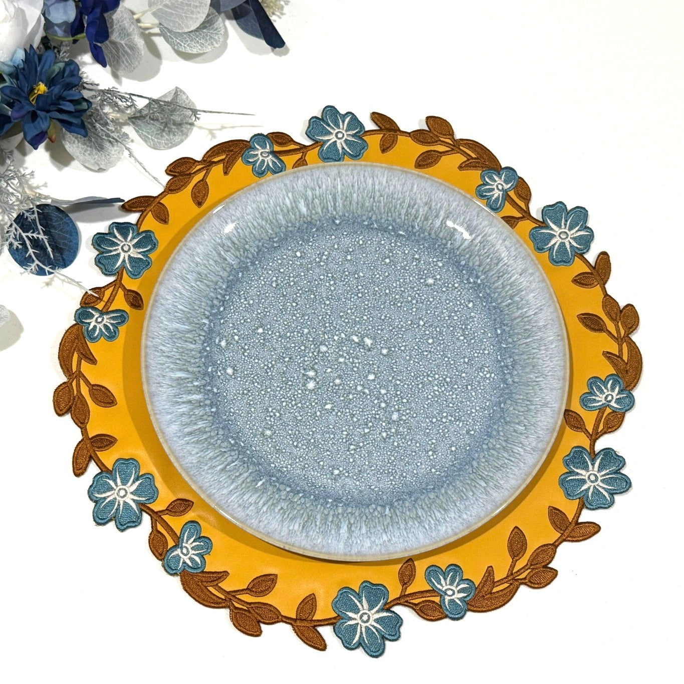 A round placemat with a yellow border and blue center, decorated with floral embroidery around the edge.