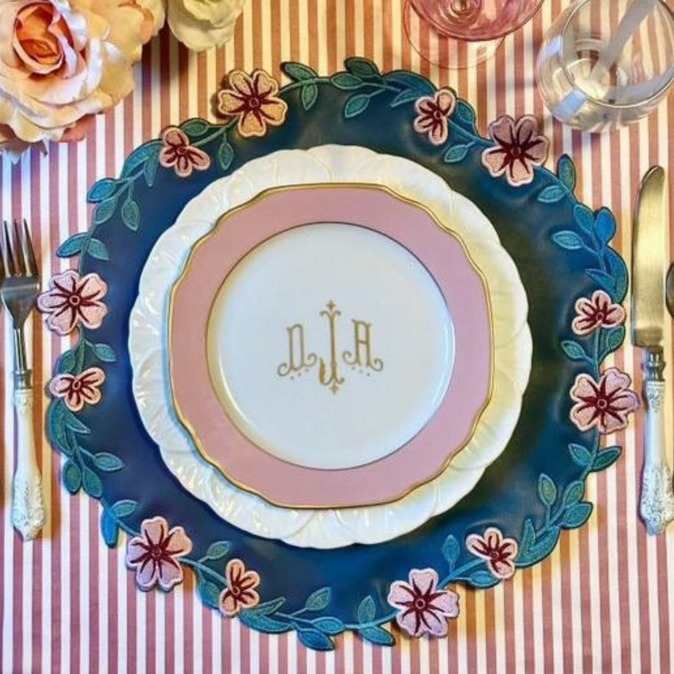 A round faux leather placemat with blue and pink floral embroidery, displayed on a table setting with cutlery and a plate.
