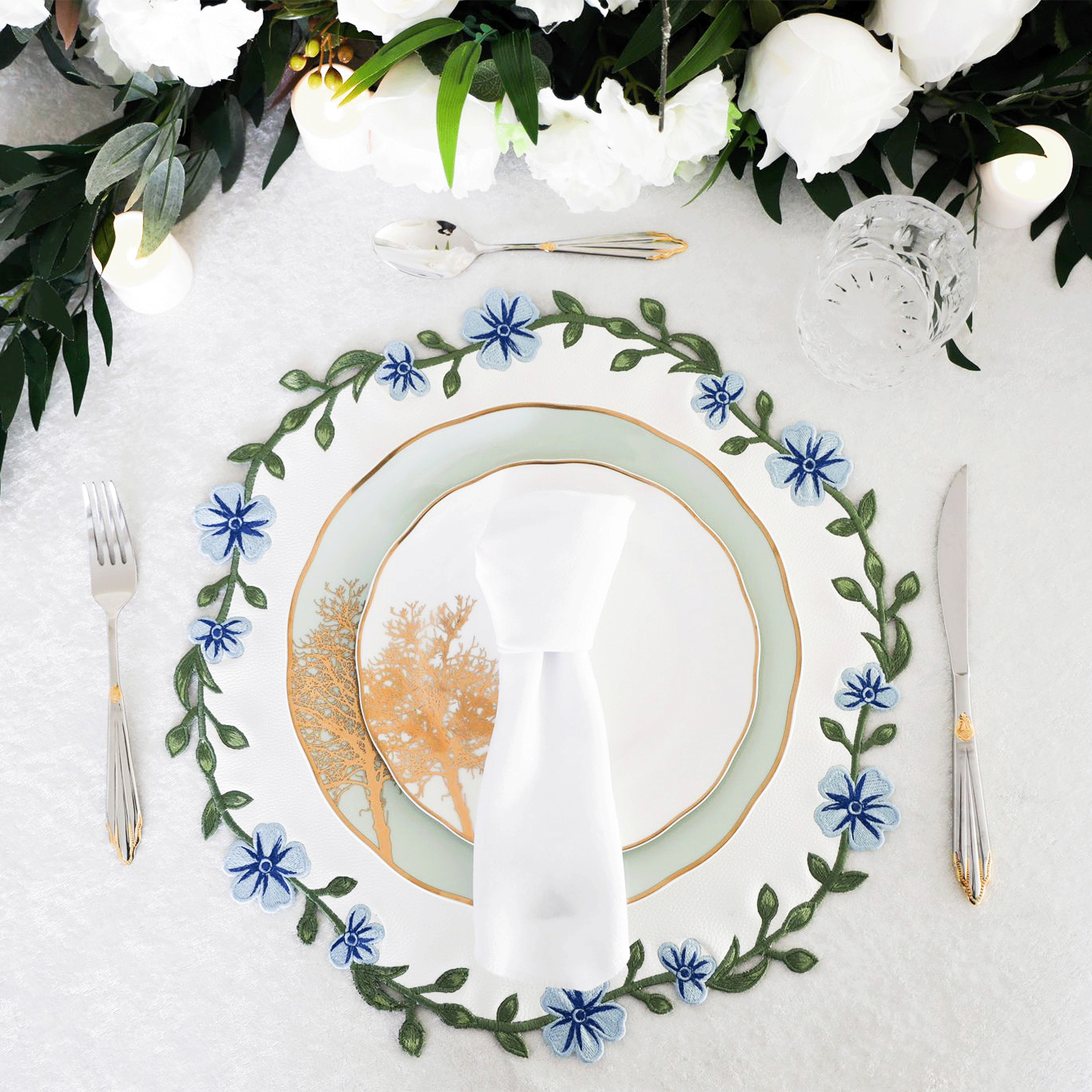 A round faux leather placemat with white and blue floral embroidery, displayed on a table setting with cutlery and a plate.