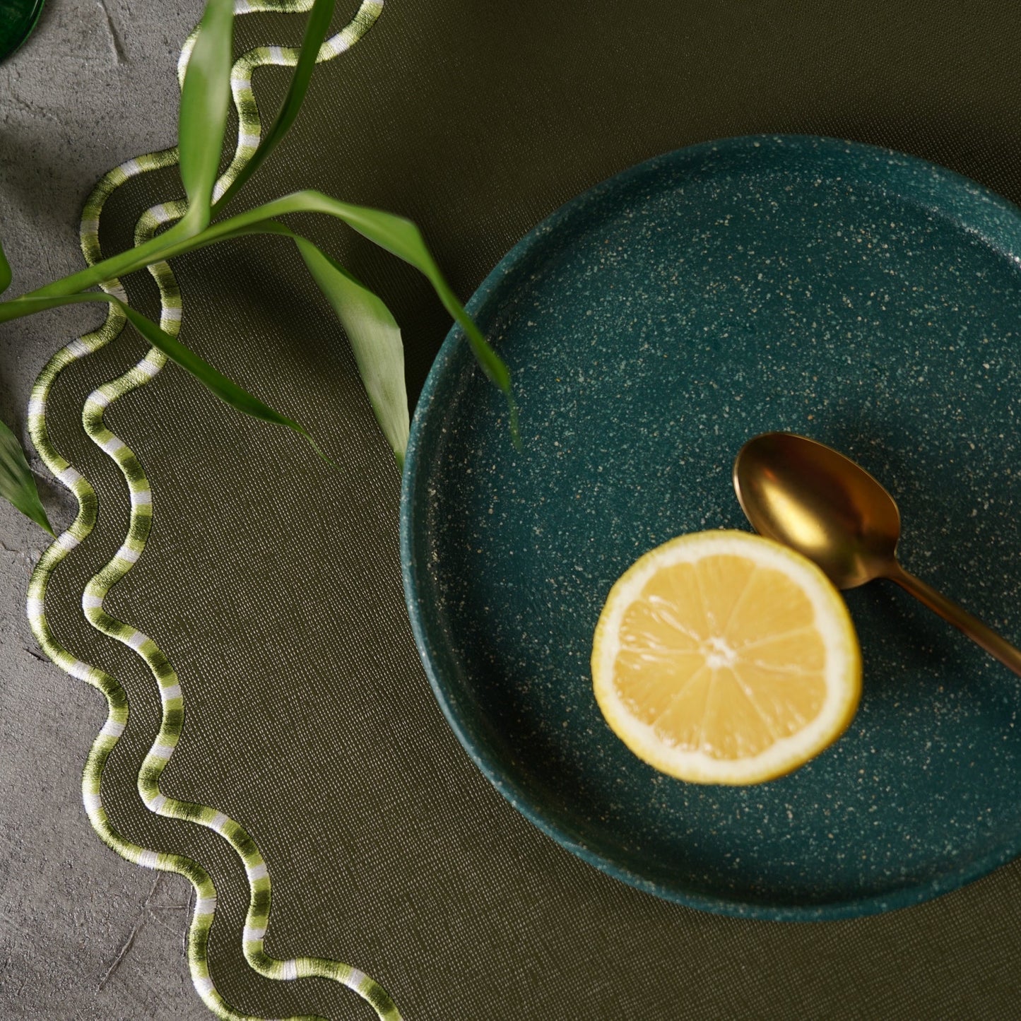 Green ceramic plate with a lemon slice and gold spoon on a dark surface with greenery.