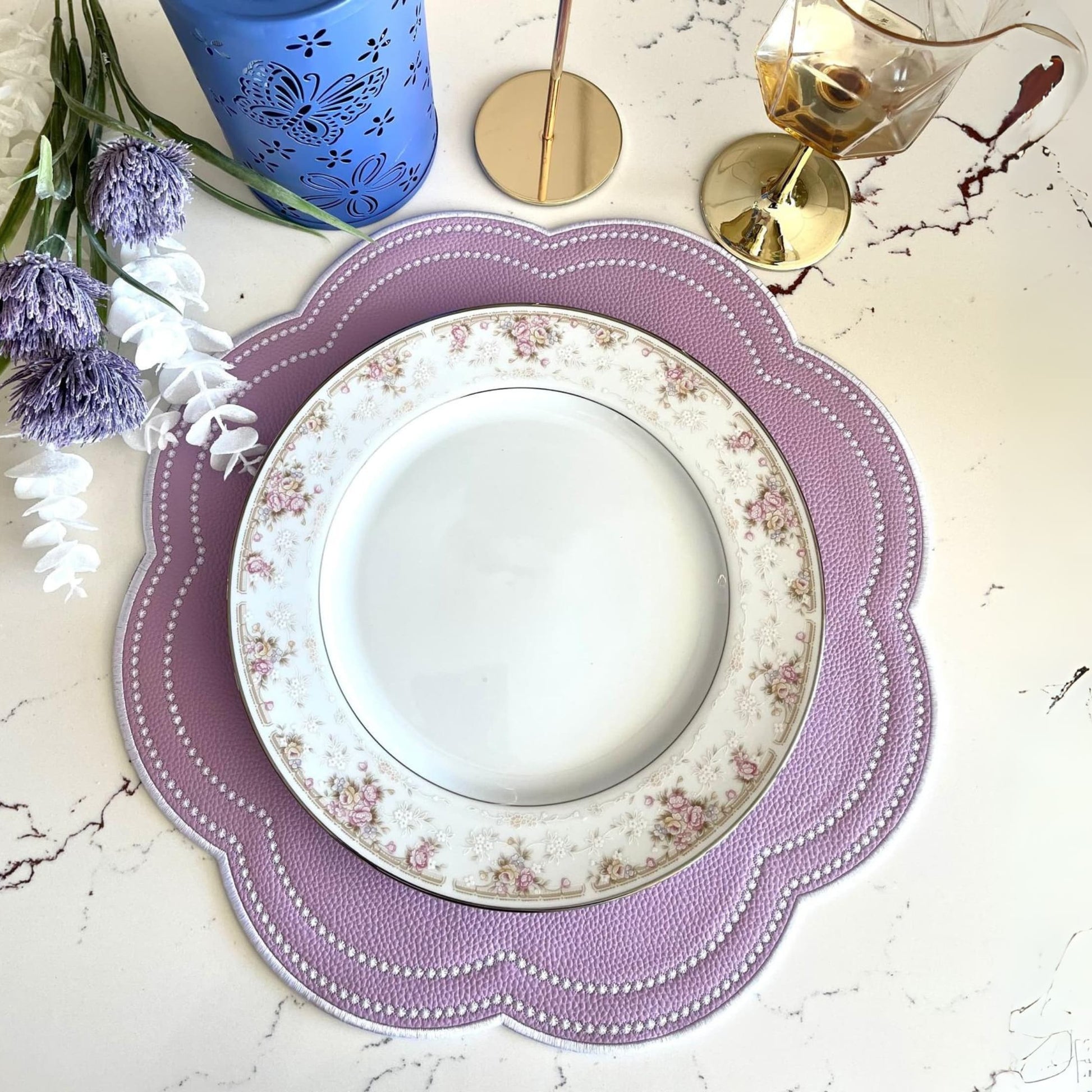 A purple faux leather placemat is laid out on a table with a white plate on top of it. There are flowers and a blue candle holder to the side, suggesting a setting for a dining table.