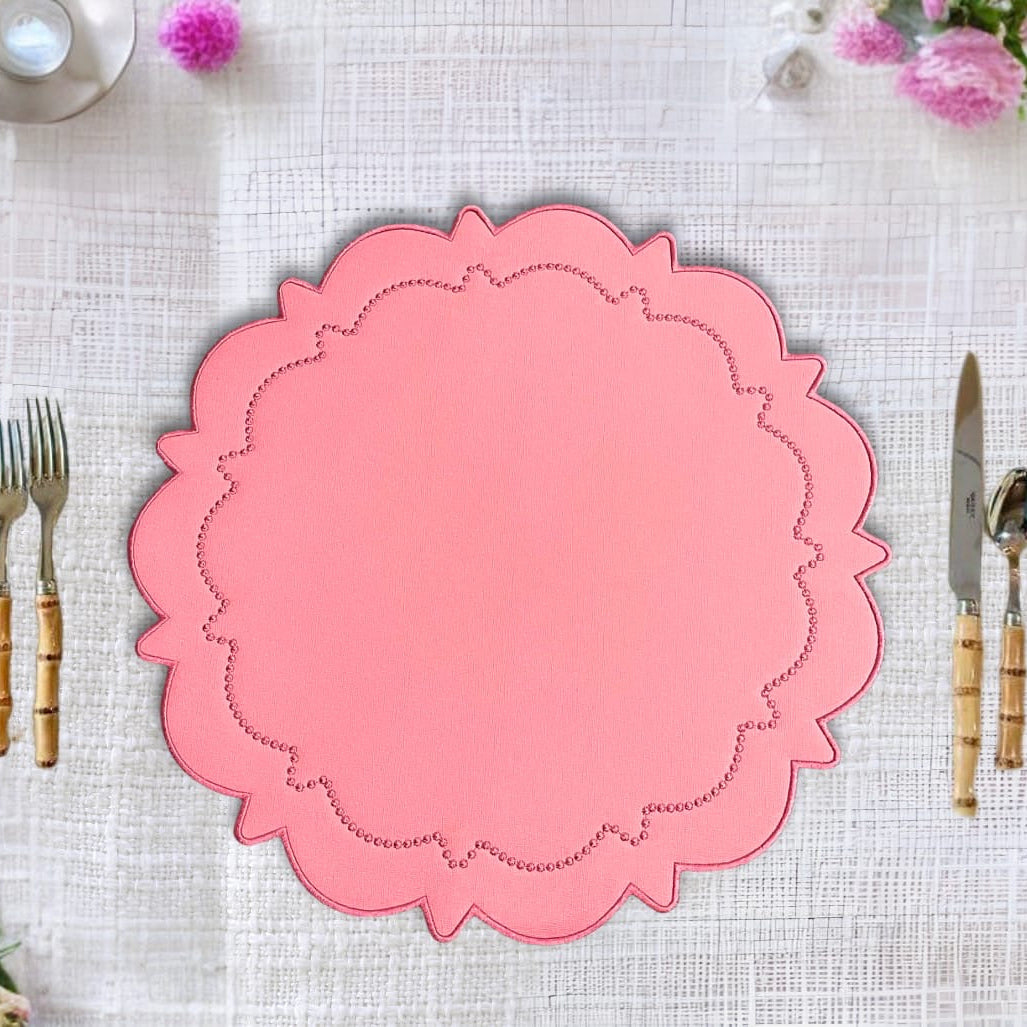 Pink placemat with decorative edge on a white tablecloth with cutlery and flowers.