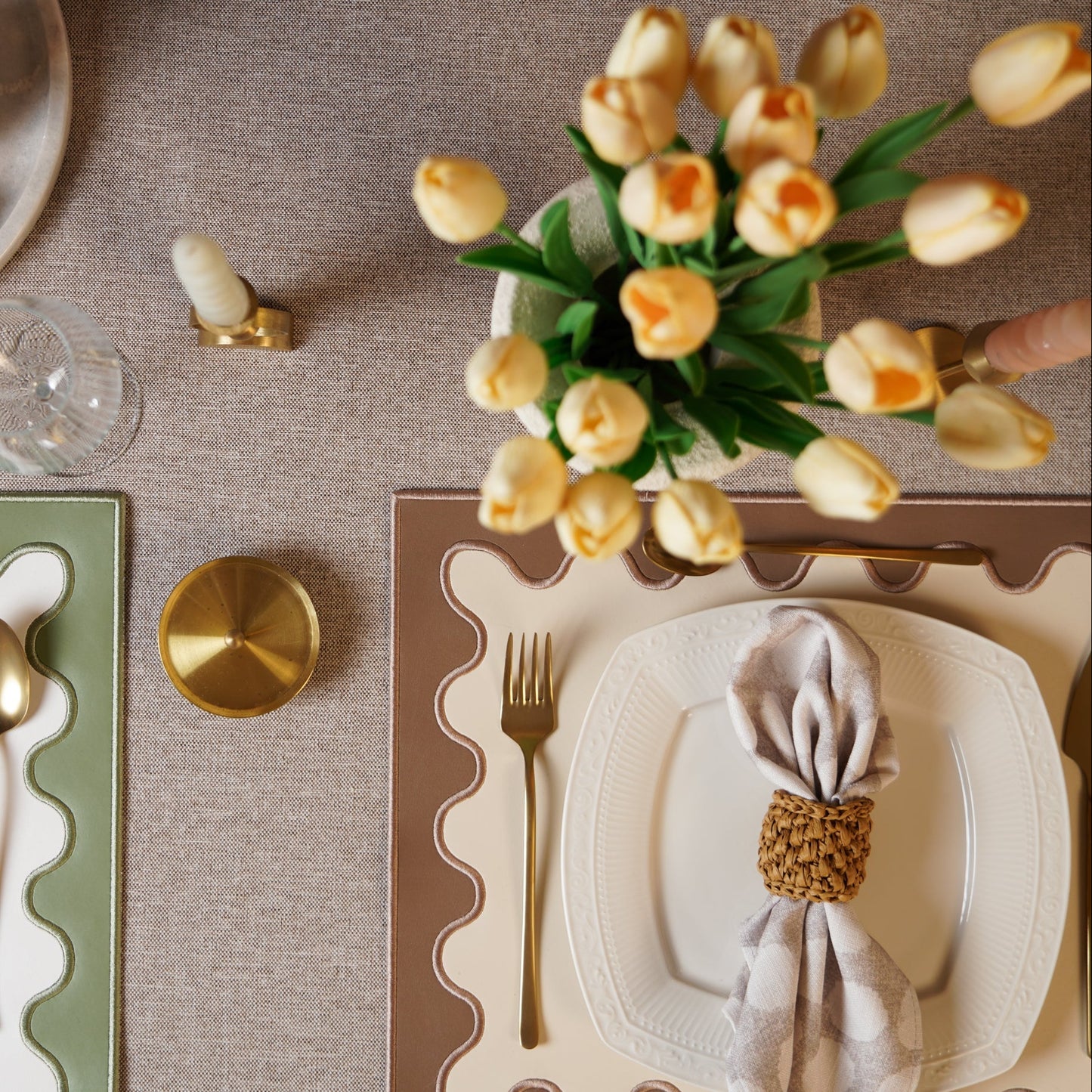 Elegant table setting with plates, napkins, cutlery, and a vase of yellow tulips on a textured surface.