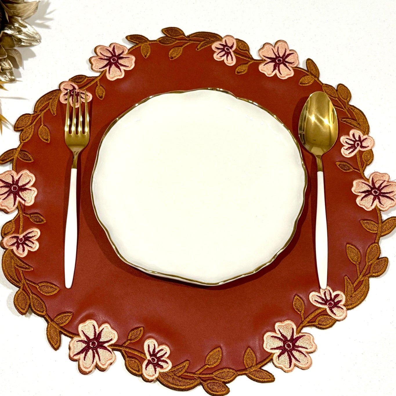 A round dark orange faux leather placemat with a floral pattern, displayed with a white plate and gold-colored utensils on a contrasting background.