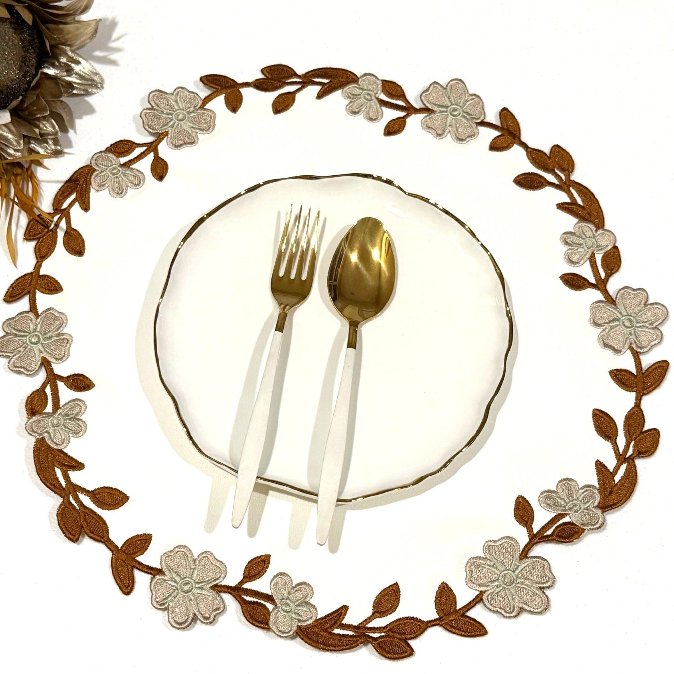 A round faux leather placemat with a white and brown floral pattern, displayed under a round white plate with gold cutlery on it. The background is white, and there is a decorative element that looks like a pine cone at the top left.