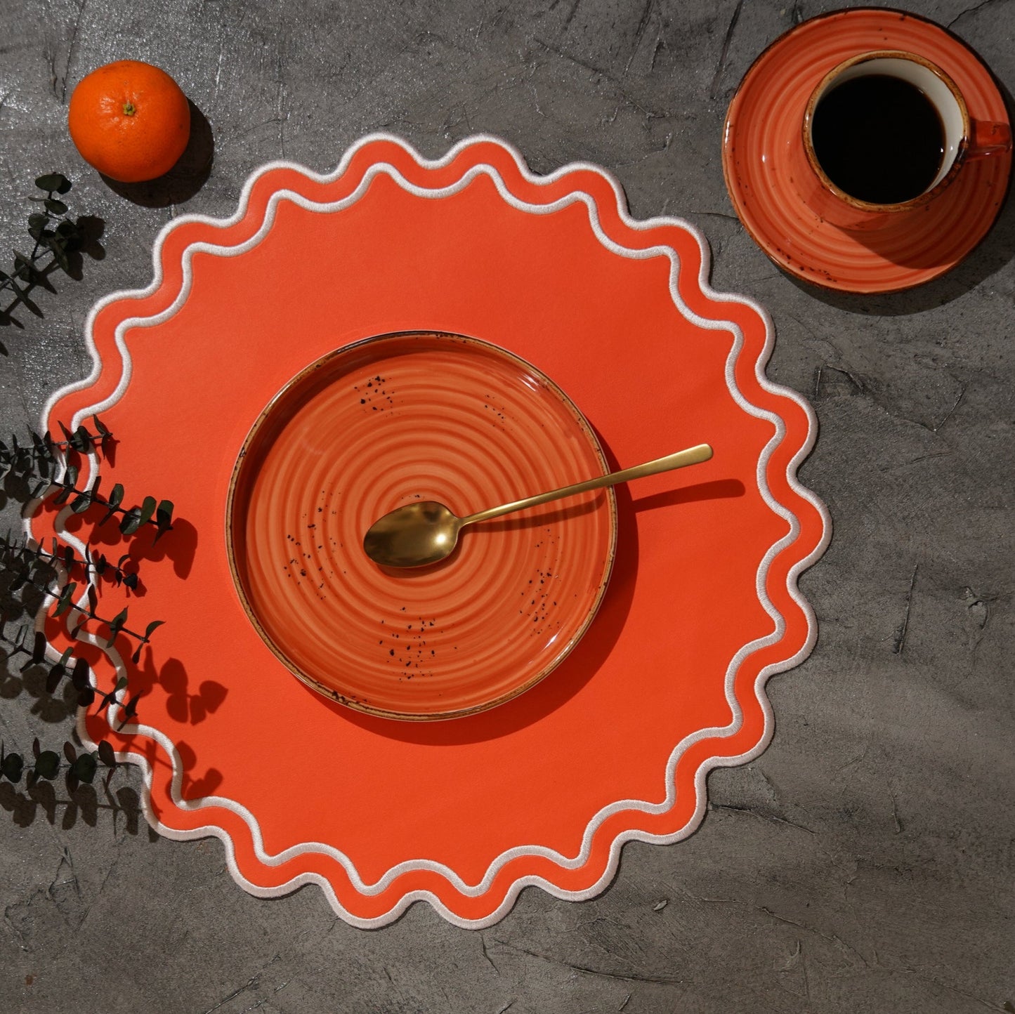 An orange faux leather placemat with a scalloped edge design, displayed on a table with a cup of coffee and an orange fruit to the side.