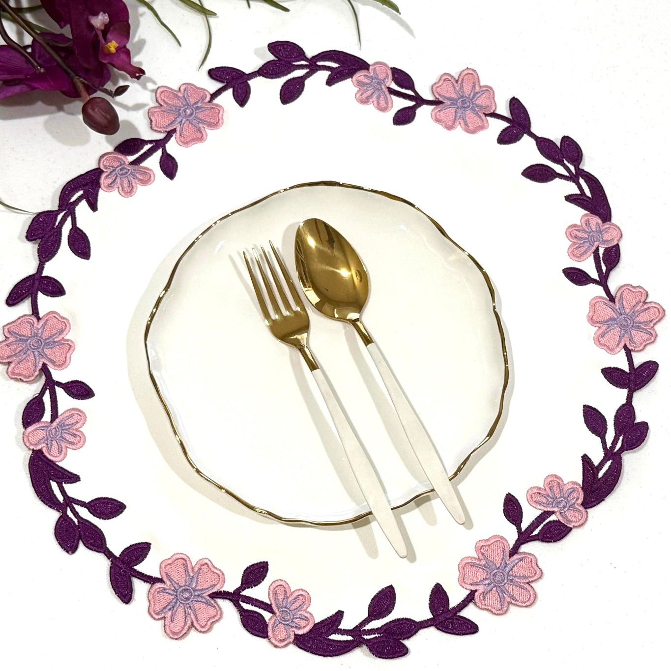 A round faux leather placemat with a floral pattern in purple and pink around a white edge, displayed under a setting of cutlery.