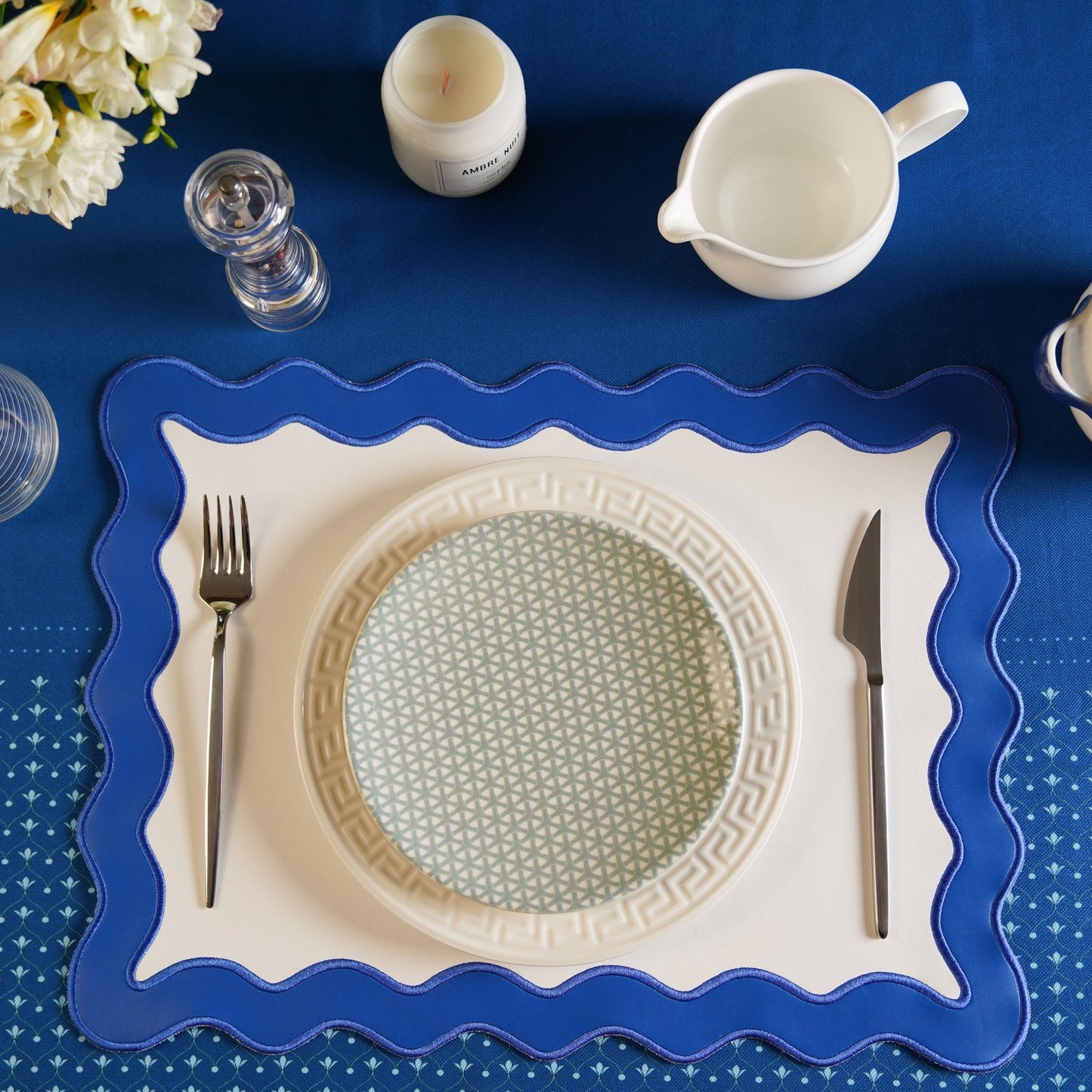 Dining table setting with a blue placemat, white plate with decorative edge, silverware, and candles on a blue tablecloth.
