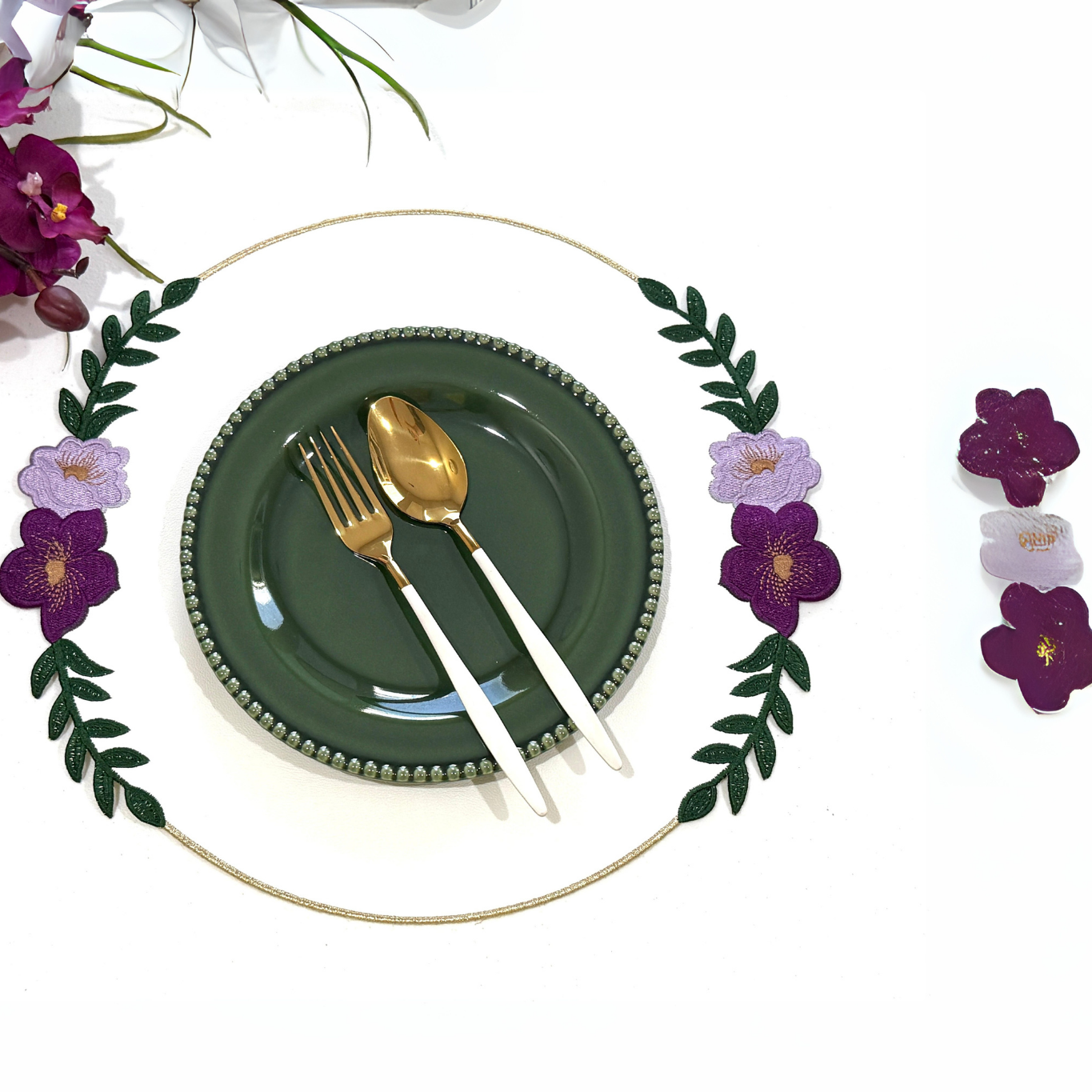 A set table with a green plate, silver cutlery, and a white faux leather placemat with embroidered details. There are purple flowers at the top of the image and decorative elements around the placemat.