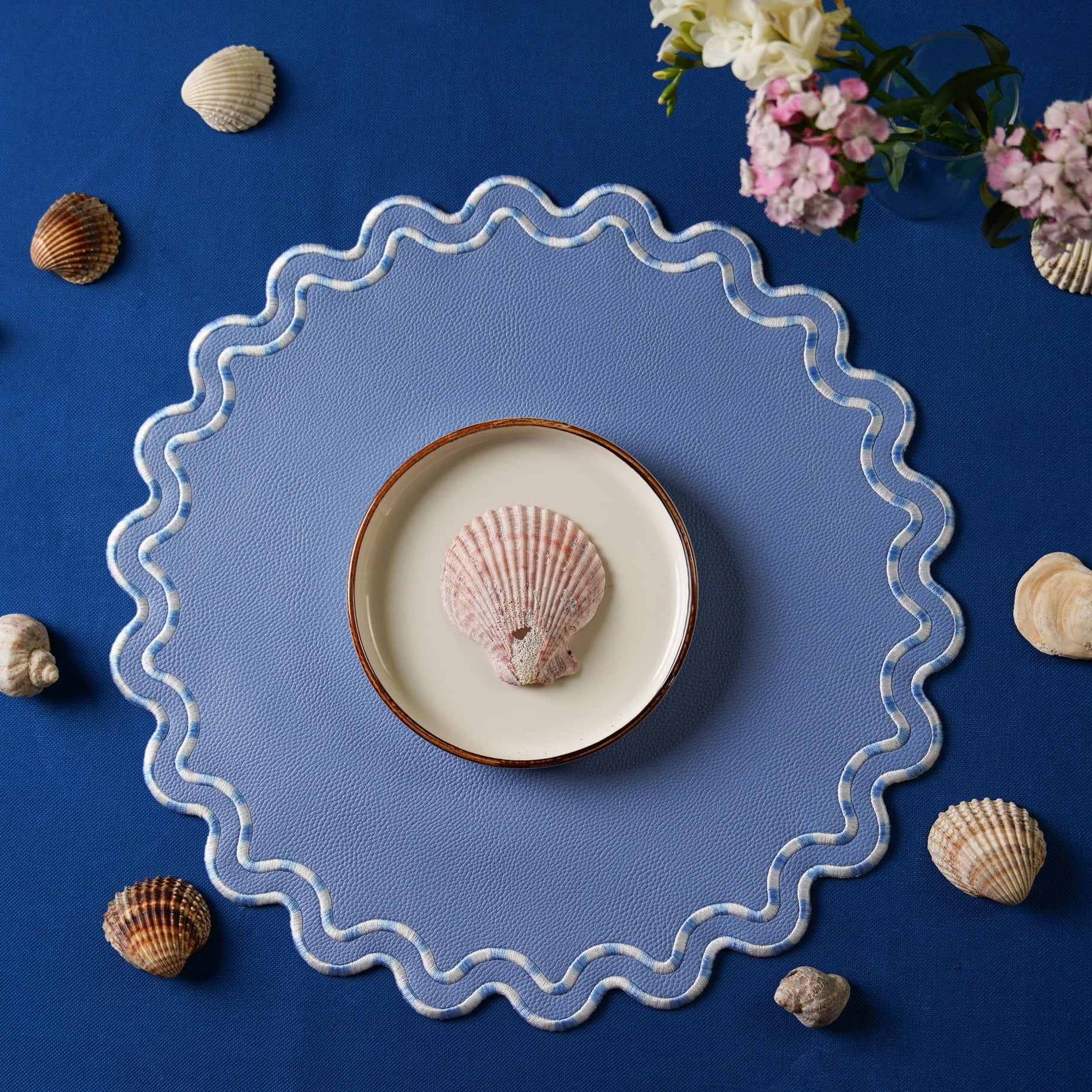 A round, light blue faux leather placemat with a scalloped edge design, displayed on a table with seashells around it.
