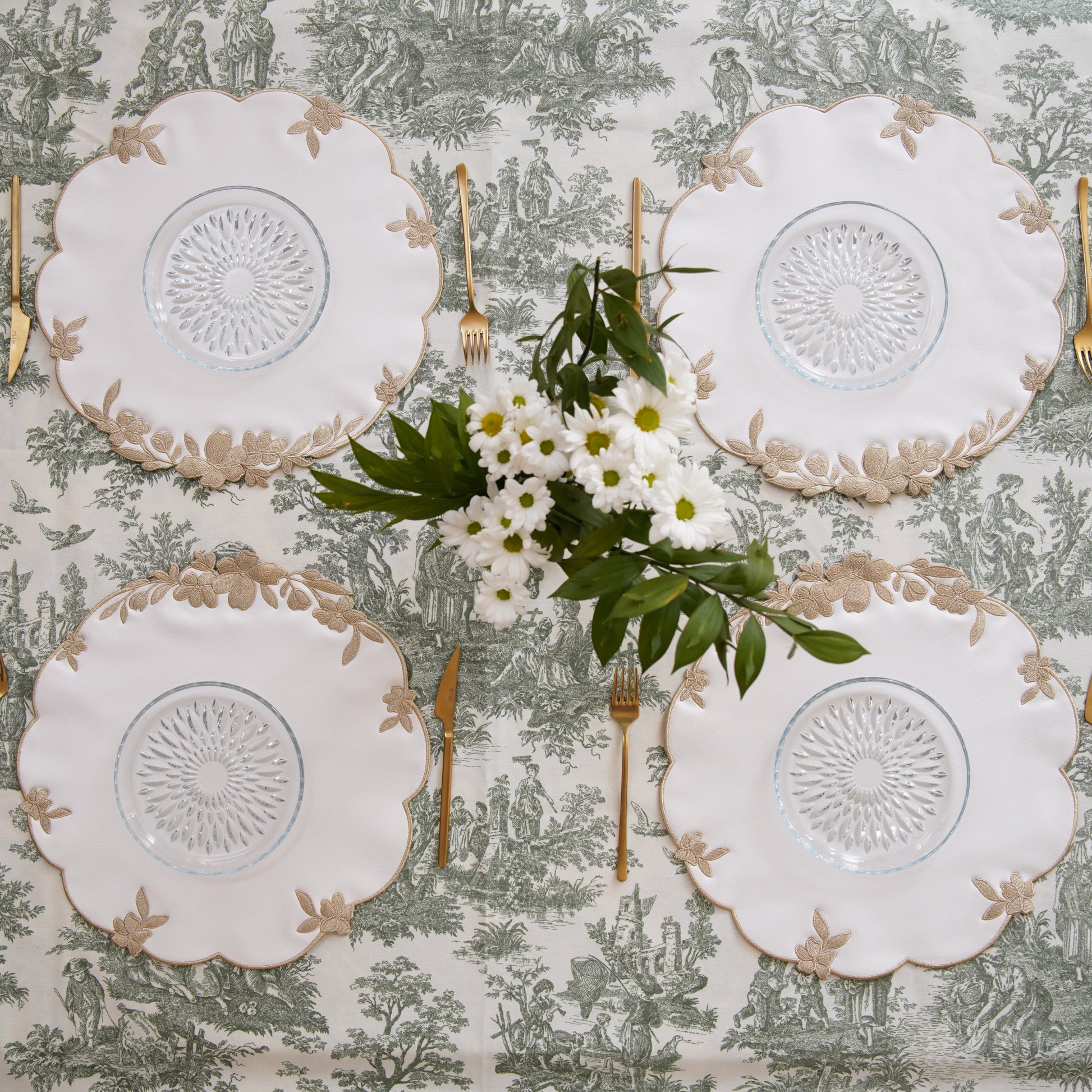 Set of four white doilies on a patterned tablecloth with silverware and flowers.