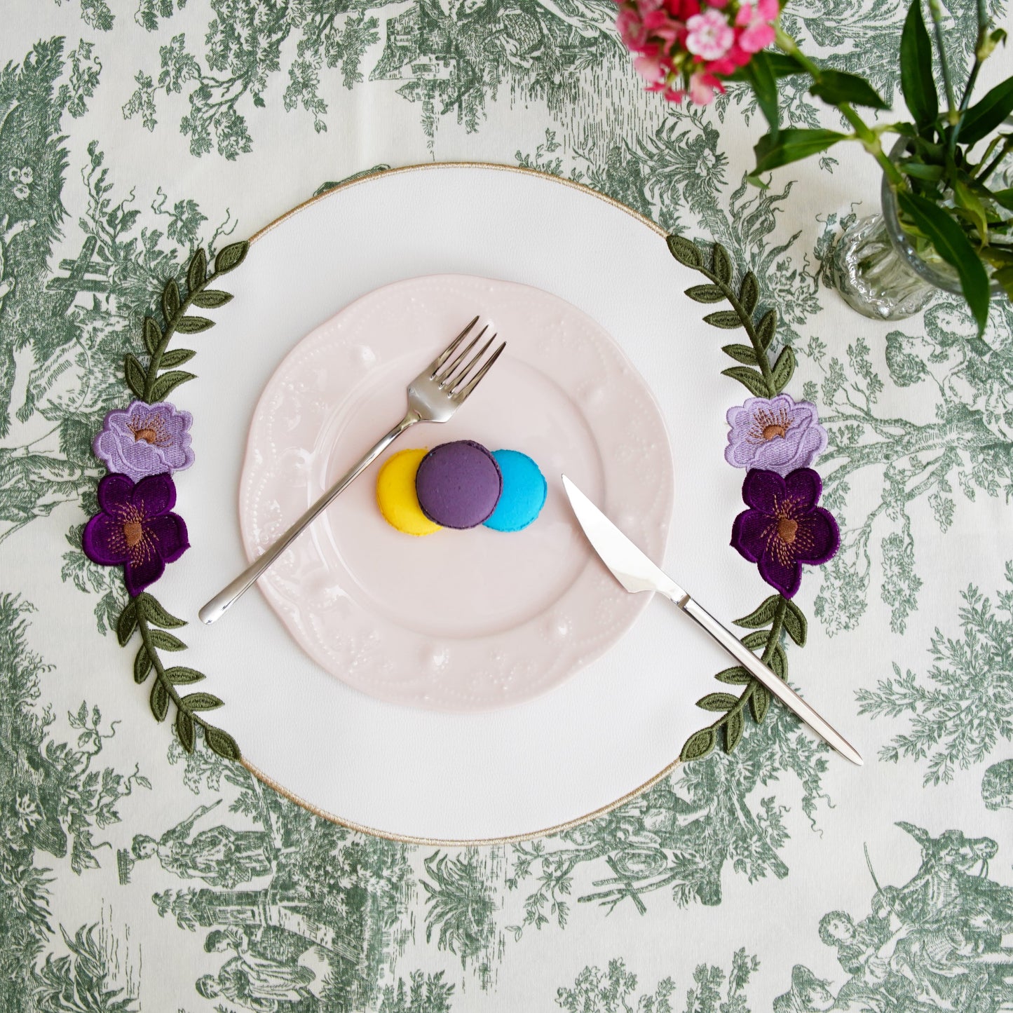 Decorative table setting with a plate, cutlery, and flowers on a patterned tablecloth.
