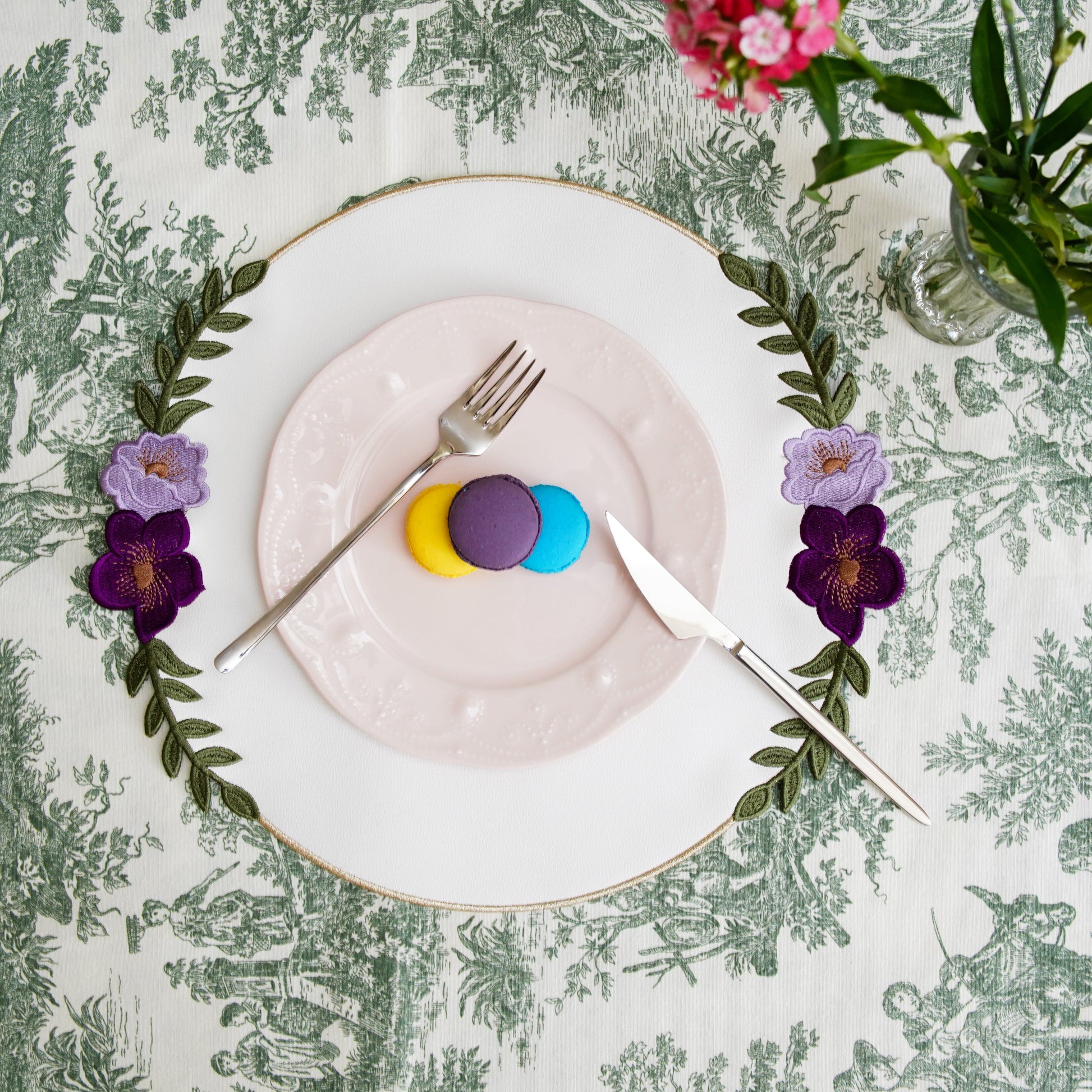 Decorative table setting with a plate, cutlery, and flowers on a patterned tablecloth.