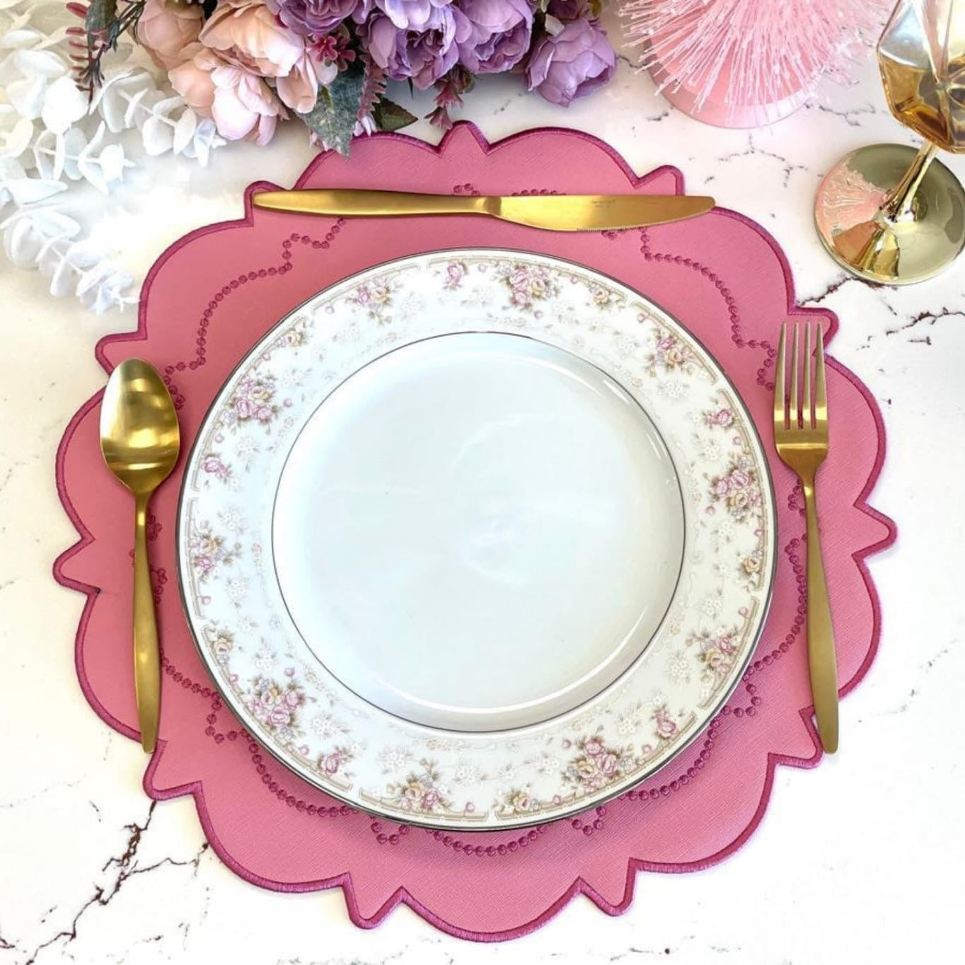 A pink faux leather placemat with a decorative edge, presented on a table setting alongside a white plate and gold cutlery.