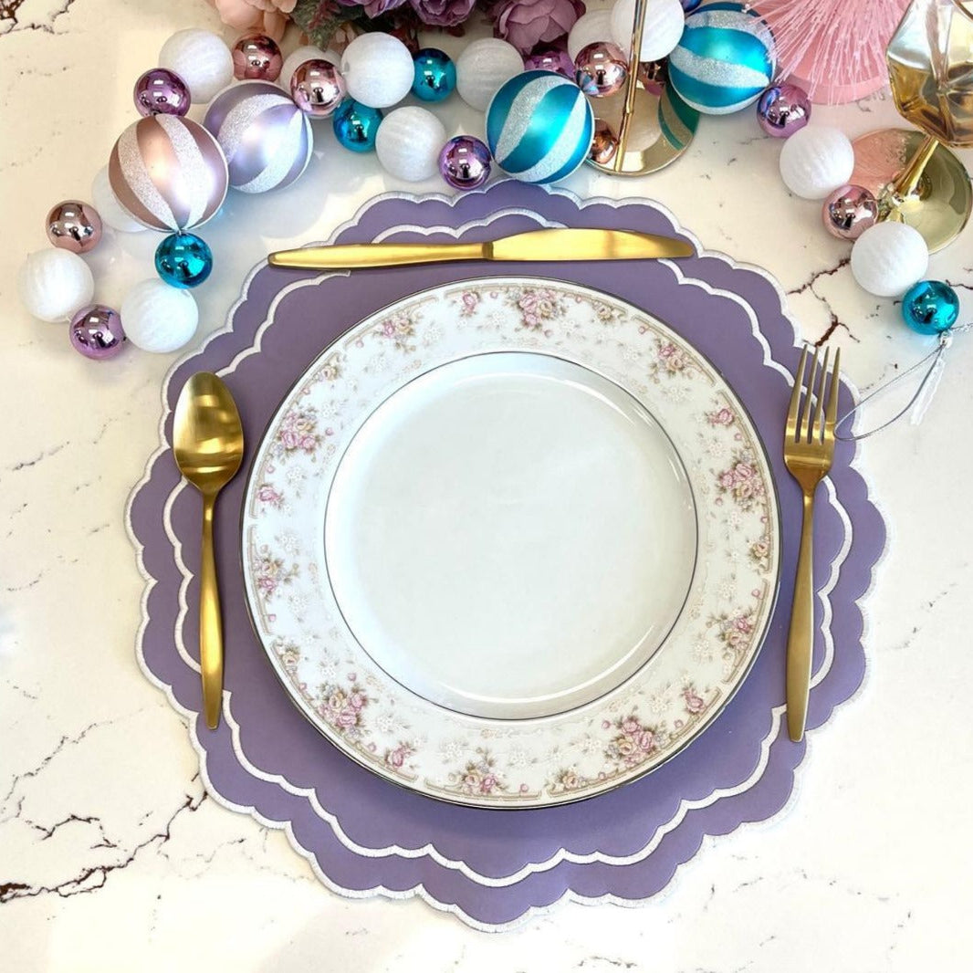 A set table with a white and scarlet faux leather placemat, a white plate, and gold cutlery. Decorative balls in various colors are also visible in the background.