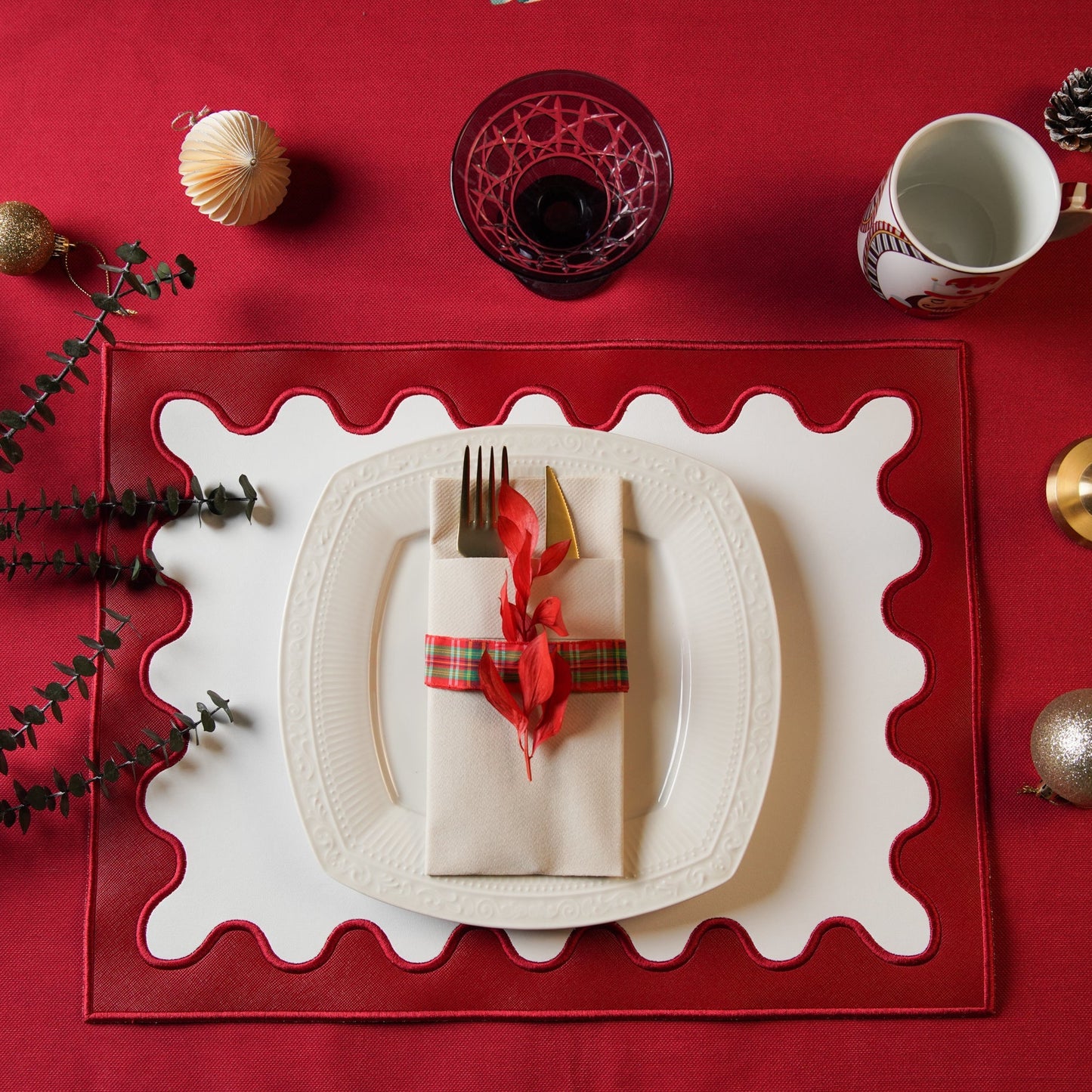 A red faux leather placemat with white embroidered detailing, presented on a table setting with a white plate, red cutlery, and decorative elements.