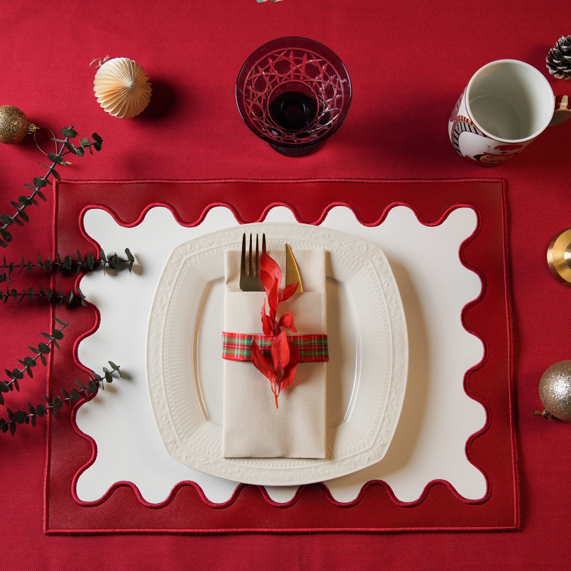 A red faux leather placemat with white embroidered detailing, presented on a table setting with a white plate, red cutlery, and decorative elements.