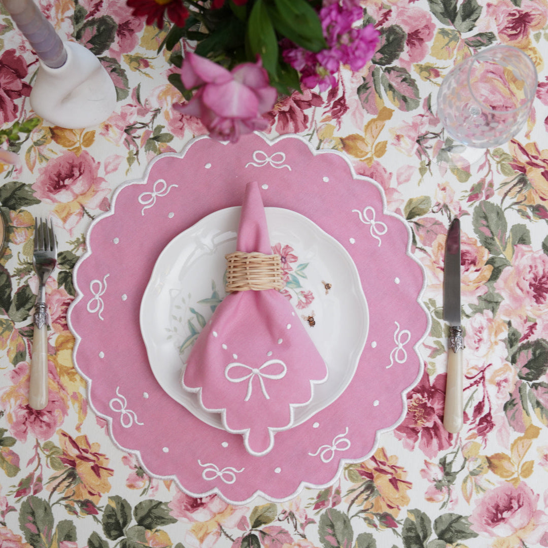 A set of pink fabric placemats with white trim, displayed on a table with floral tablecloth, along with a napkin tied with a ribbon and cutlery.
