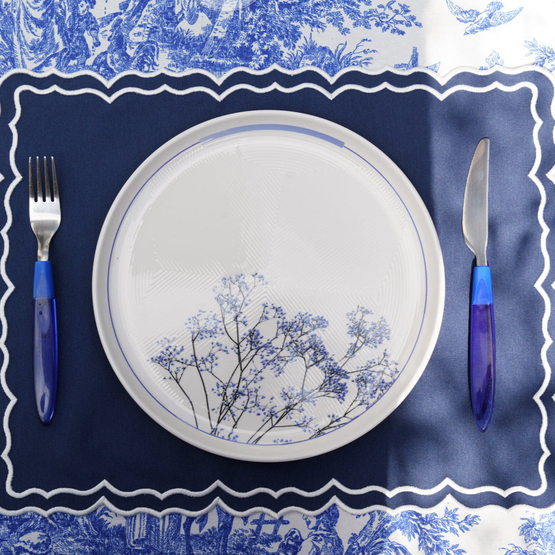 A rectangular dark blue fabric placemat with white trim, displayed on a table with a plate and cutlery, with a floral patterned tablecloth in the background.