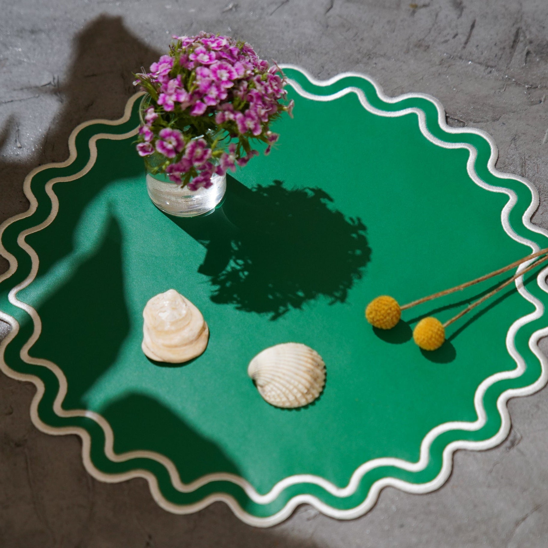 Green decorative plate with a vase of purple flowers, seashells, and yellow flowers on a gray surface.