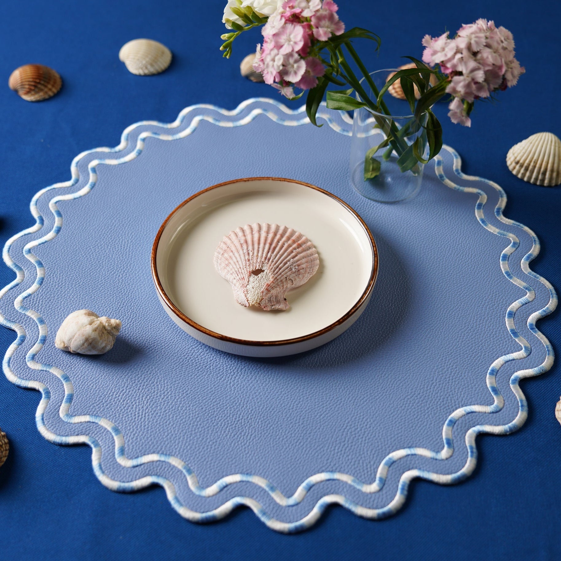 Decorative placemat with scalloped edge on a blue surface, featuring a bowl with a shell and flowers.