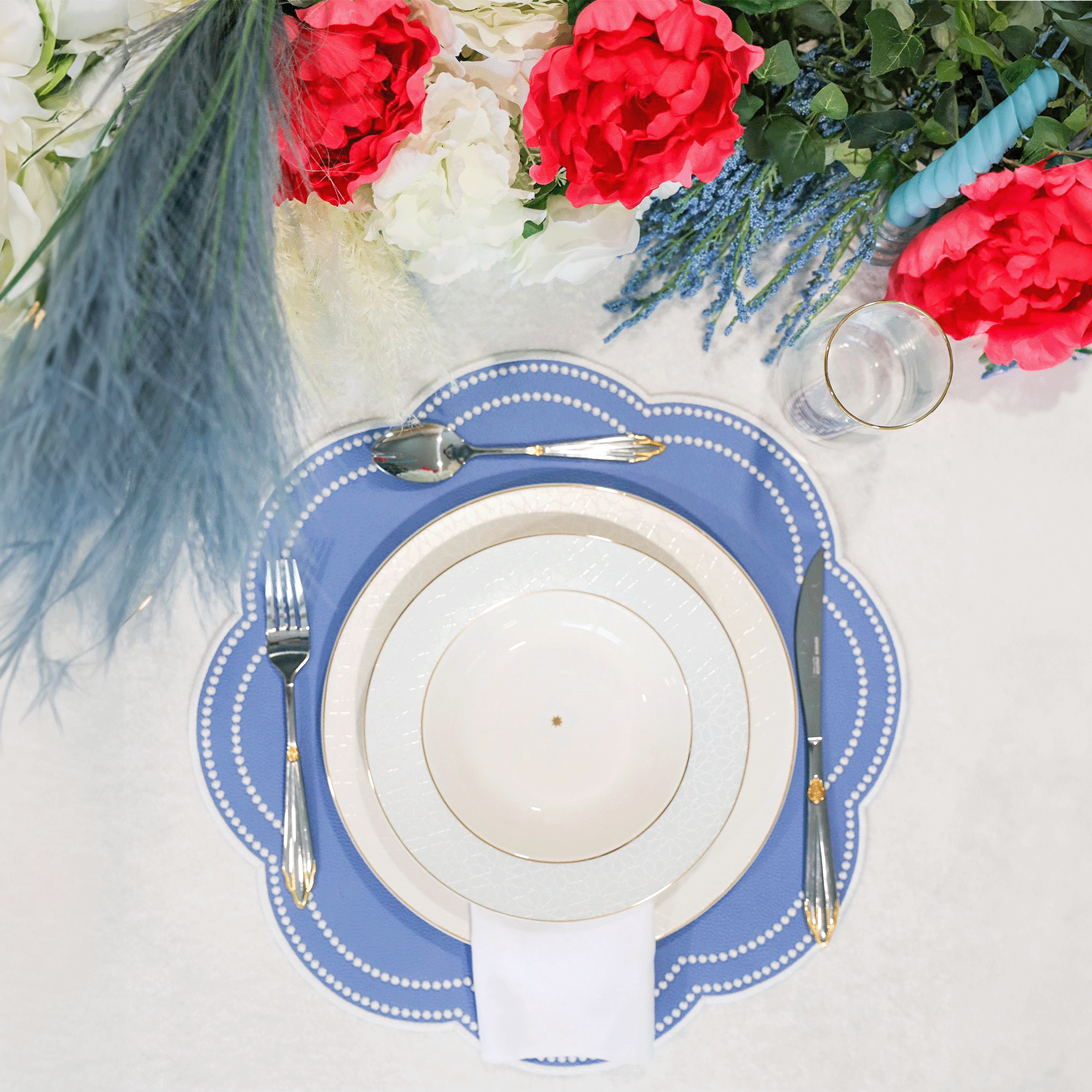 A set dining table with a light blue faux leather placemat, white plate, and silverware. The table is adorned with a red and white floral arrangement and has a grey stone-like appearance.