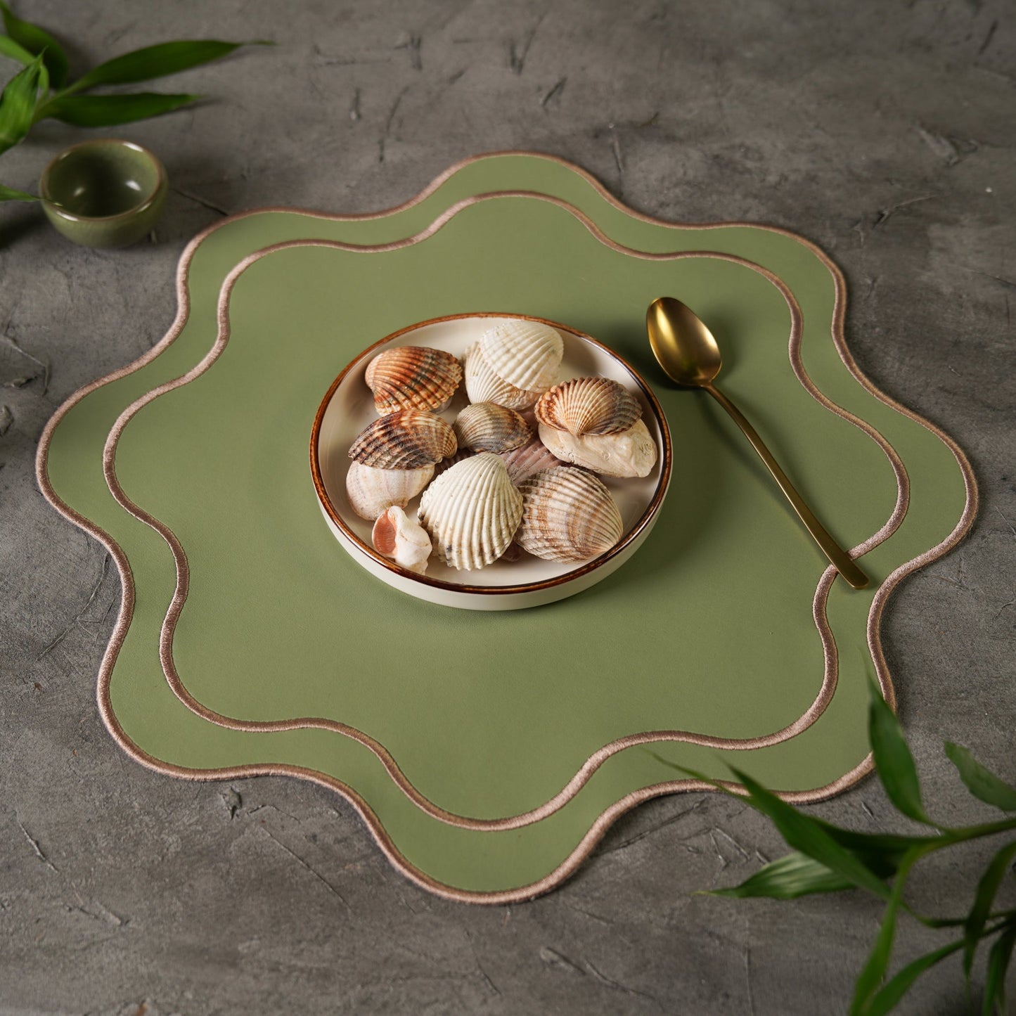 Green ceramic plate with scalloped edge holding seashells on a gray surface with greenery.