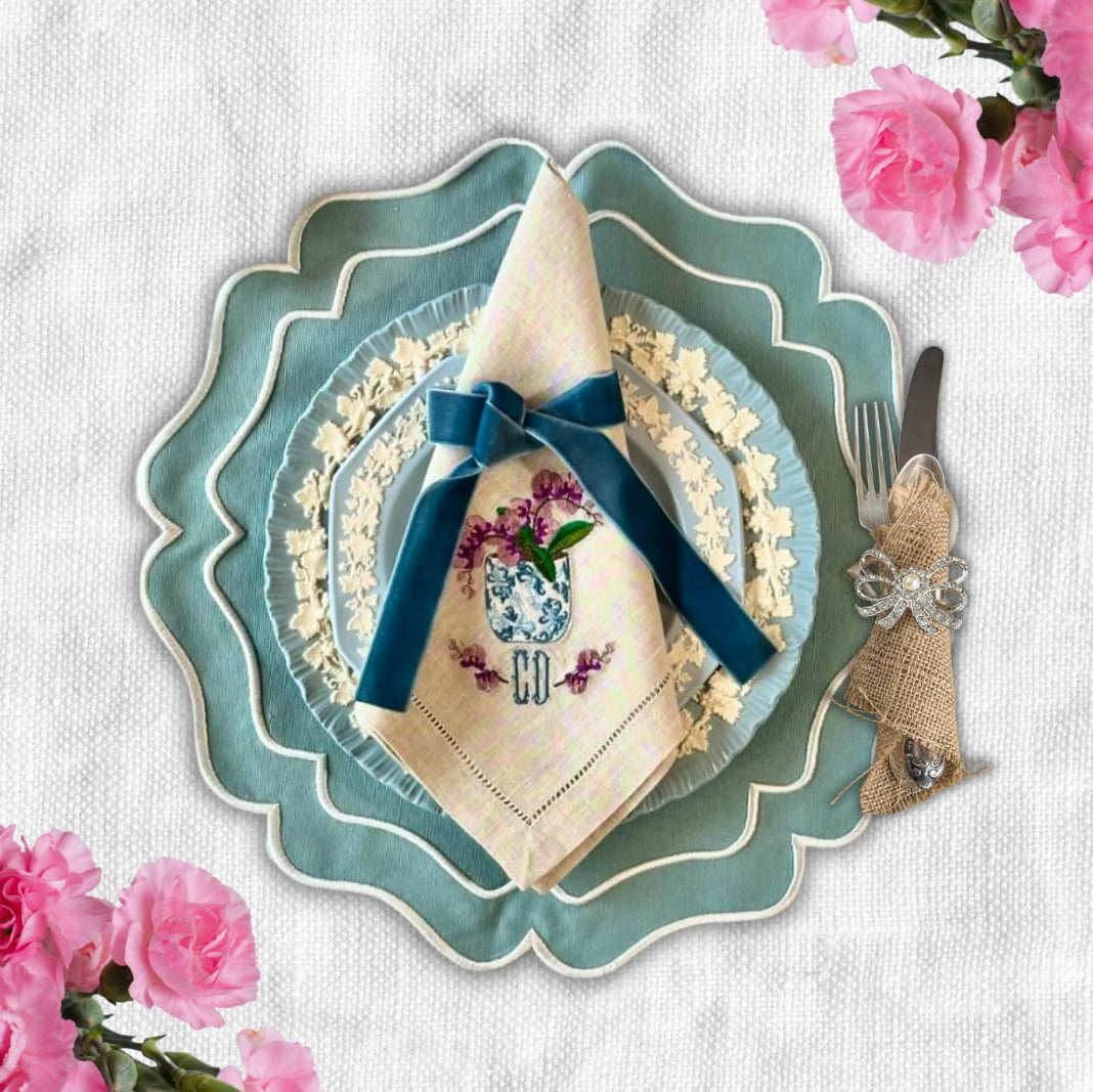 A set of white embroidered placemats with blue ribbons, displayed on a table with a floral pattern in the background.