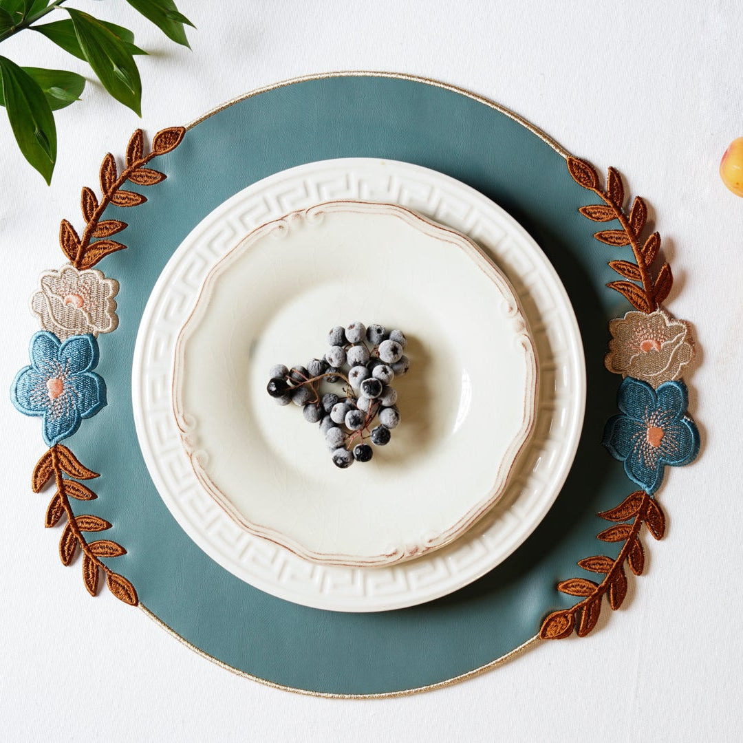 A round faux leather placemat in blue with floral embroidery, displayed on a white surface, with a white plate and decorative orange balls in the background.