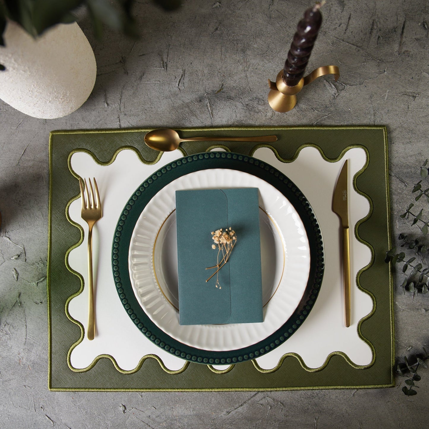 Dining table setting with green plate, white napkin, gold cutlery, and decorative elements on a textured surface.
