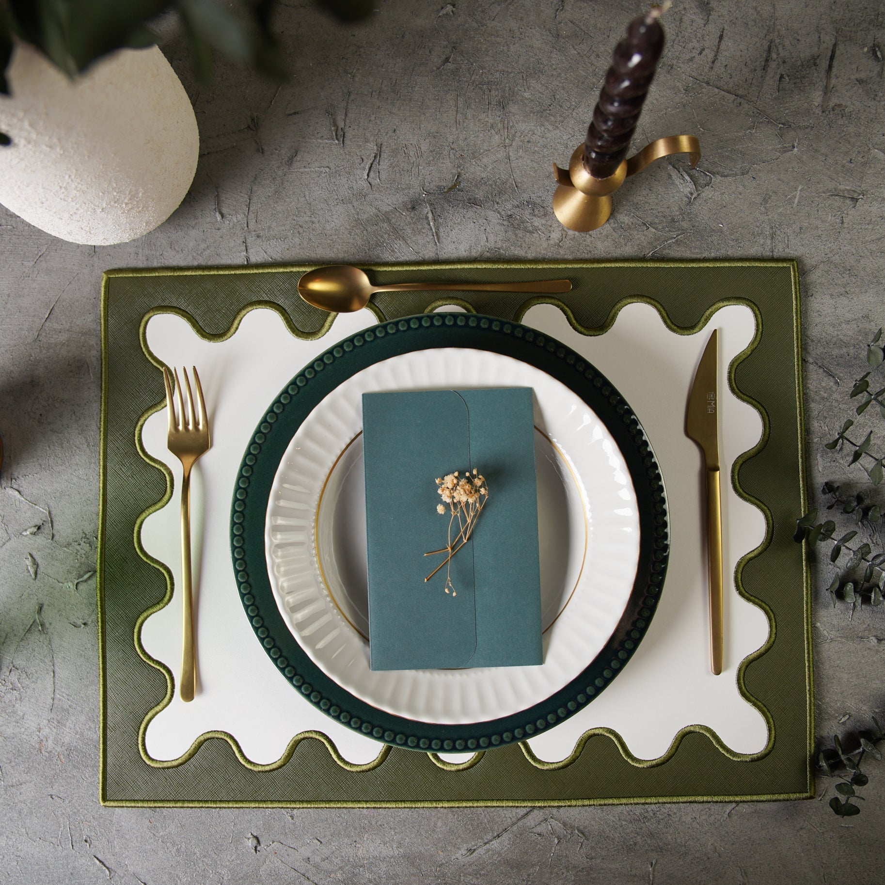 Dining table setting with green plate, white napkin, gold cutlery, and decorative elements on a textured surface.