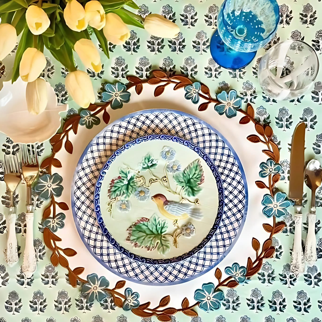 A set table with a faux leather placemat in white, blue, and brown with a floral pattern, surrounded by dishes and cutlery.