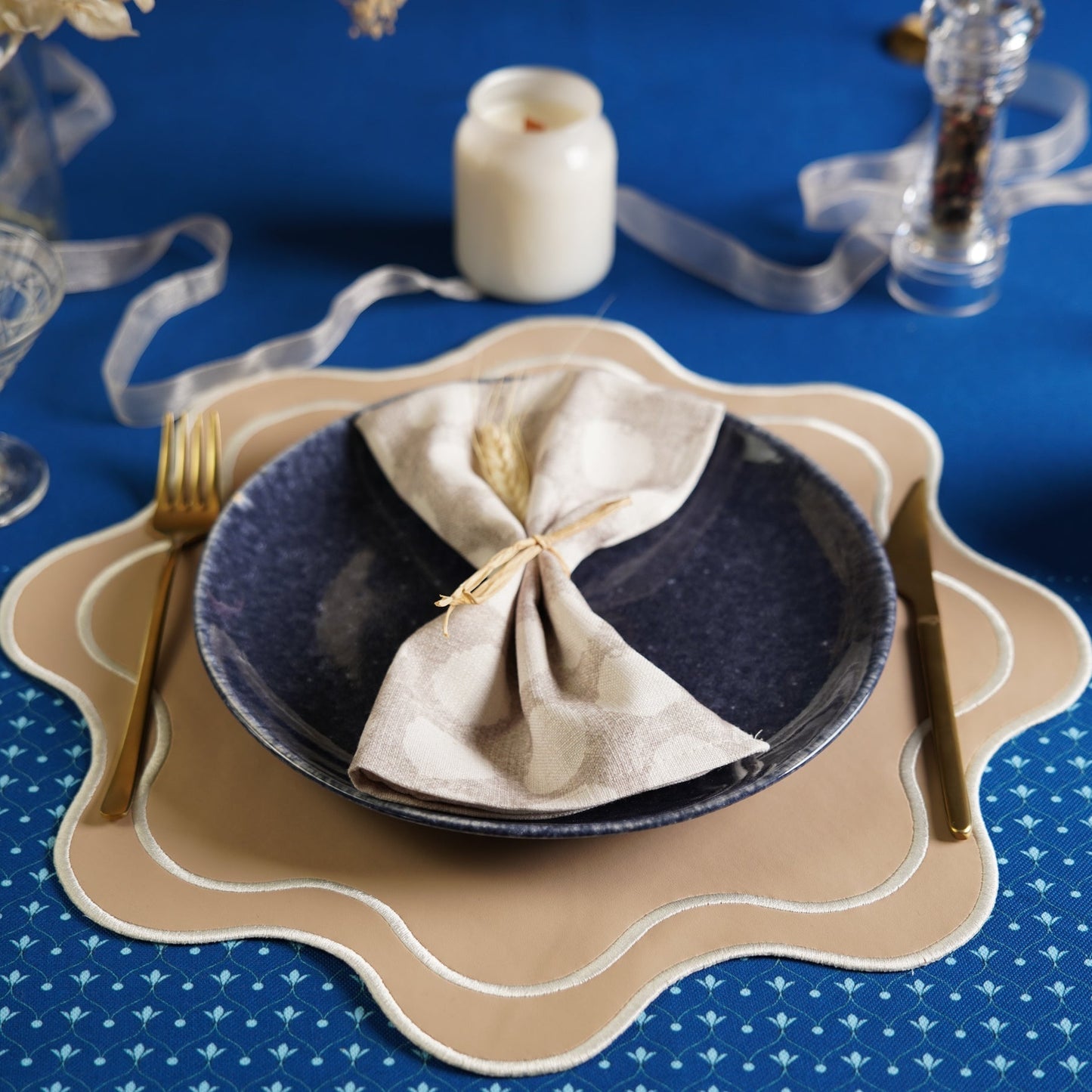 Table setting with a blue plate, beige napkin, gold fork and knife on a blue tablecloth.