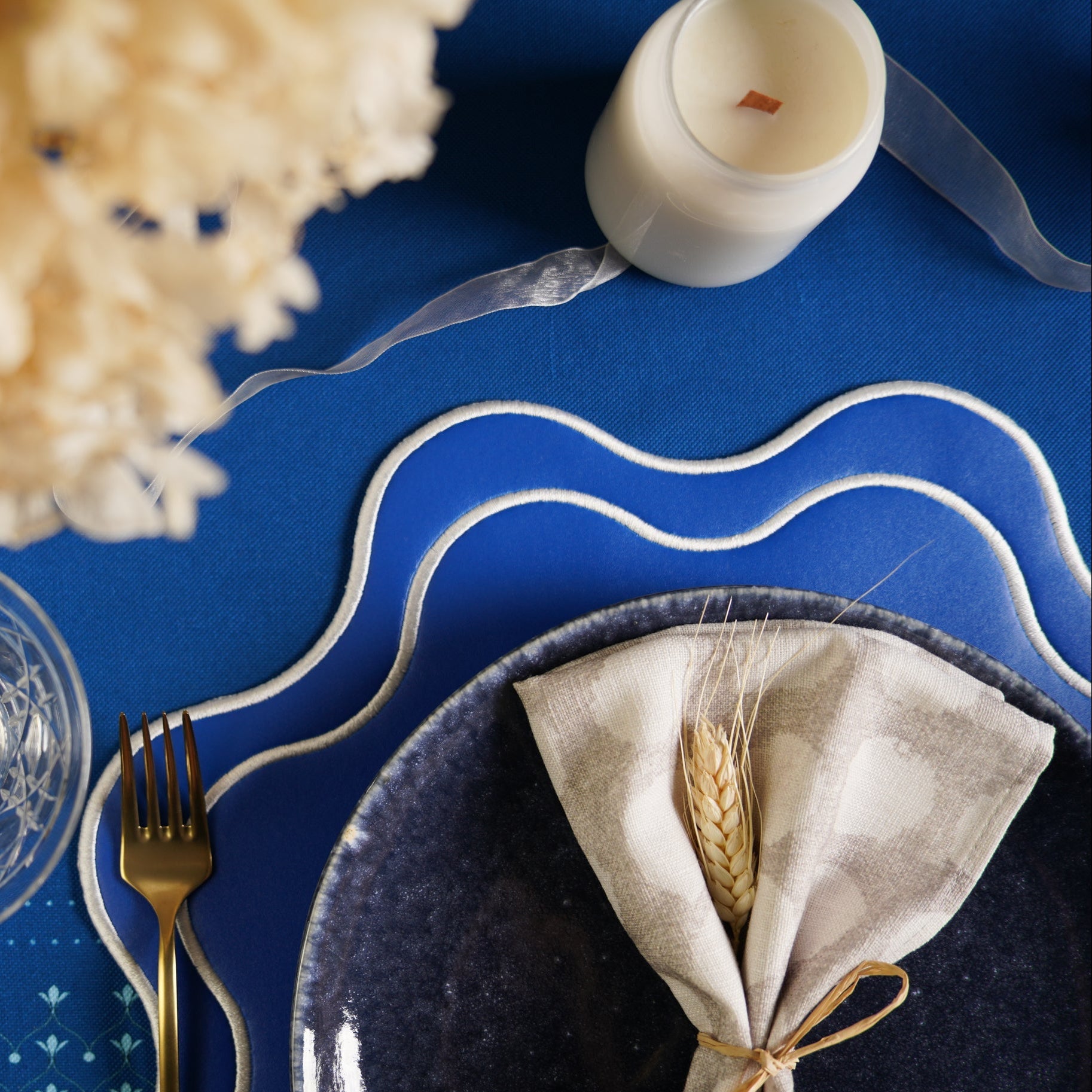 Dinner setting with a blue plate, gold fork, white napkin with wheat, and candle on a blue tablecloth.