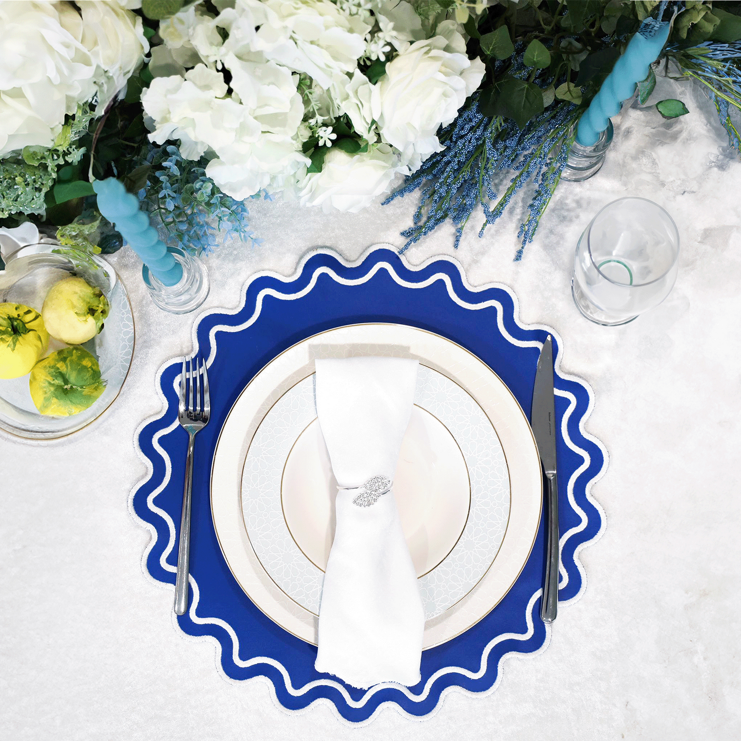 A blue faux leather placemat is laid on a white surface, featuring a round shape with a scalloped edge design. There is a white plate and silverware set on top of the placemat, and a glass with a lemon slice in it to the side.