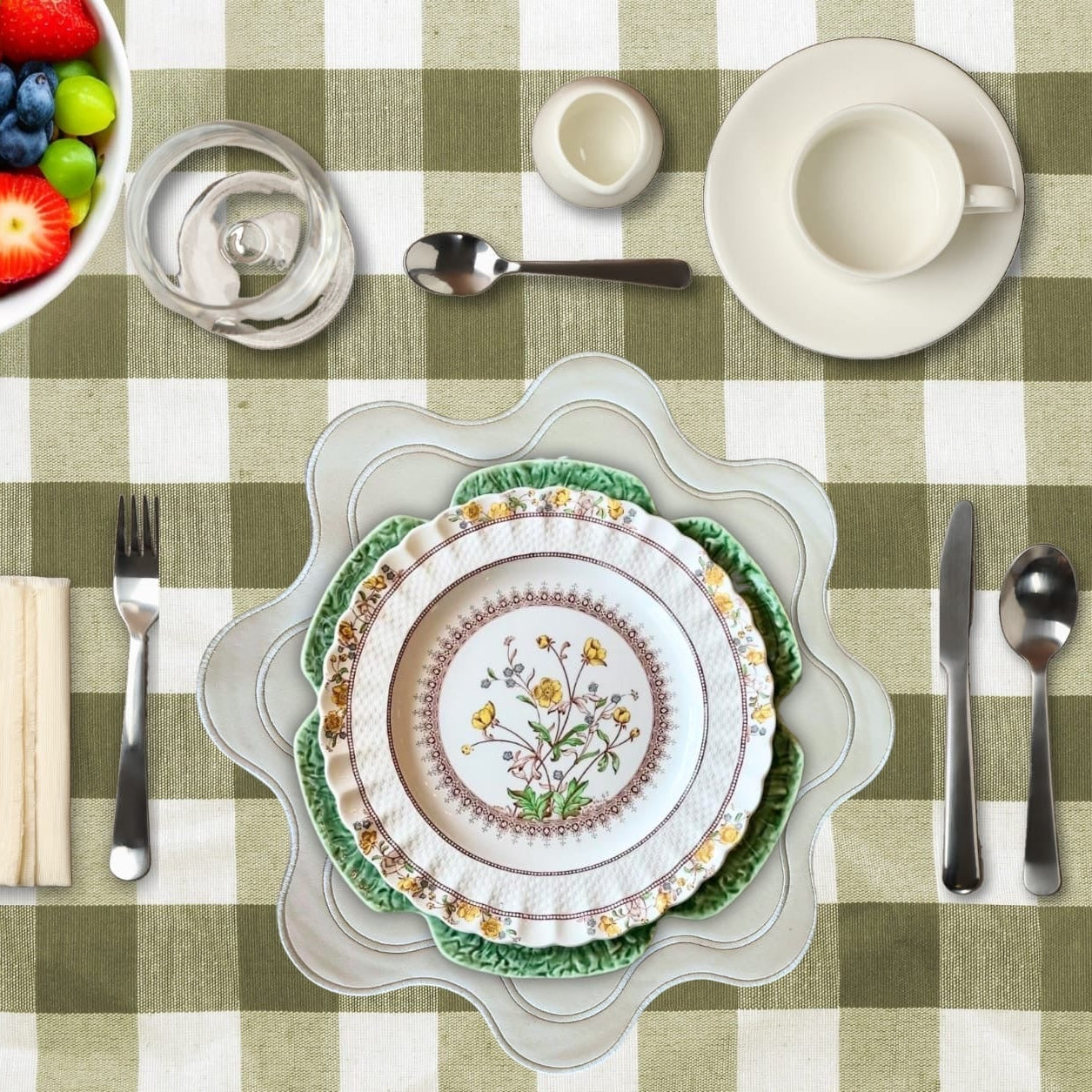 Table setting with floral plates, silverware, and a bowl of fruit on a green checkered tablecloth.