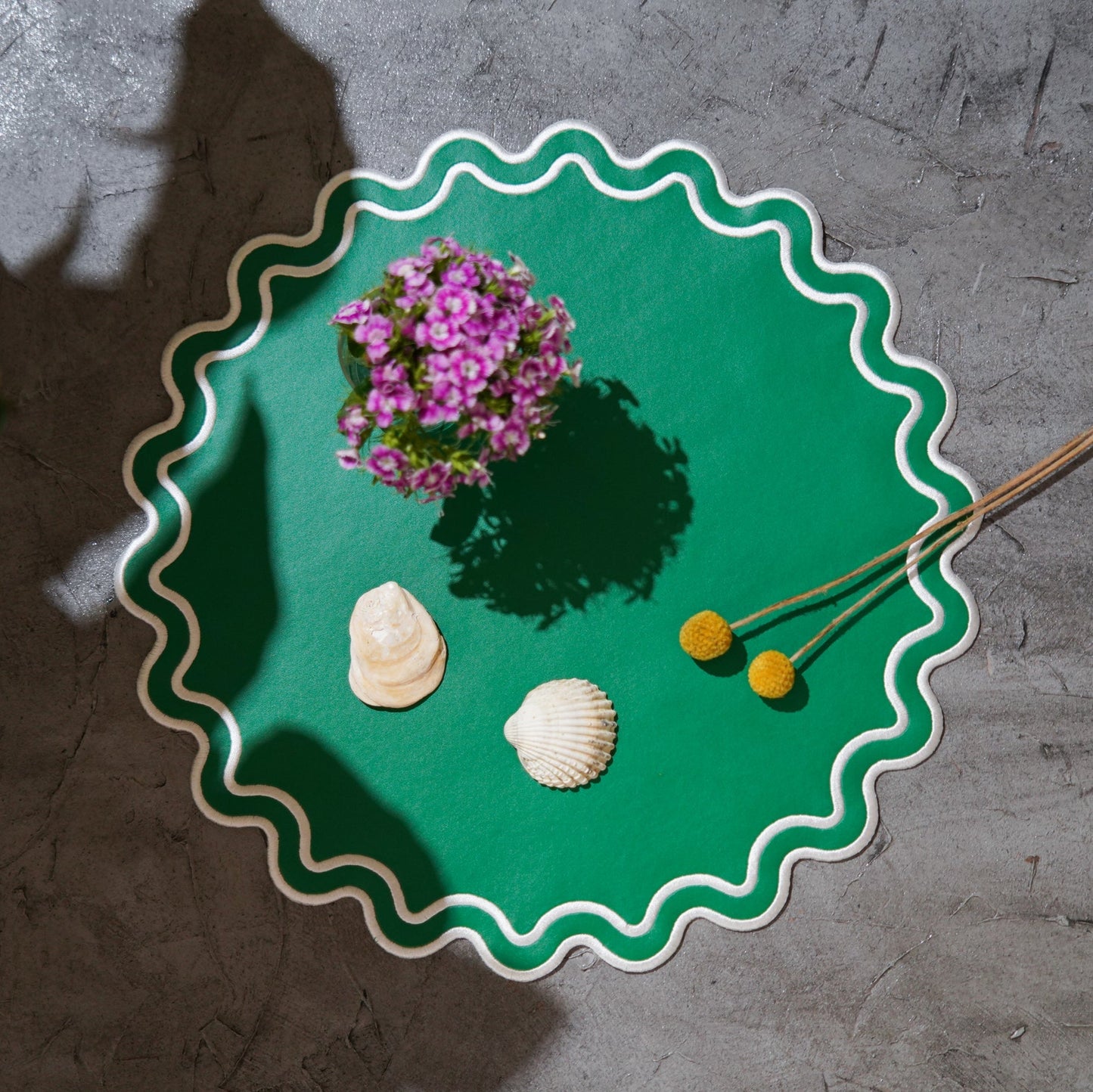 A round green faux leather placemat with a scalloped edge design, displayed on a table with a vase of flowers and decorative seashells.