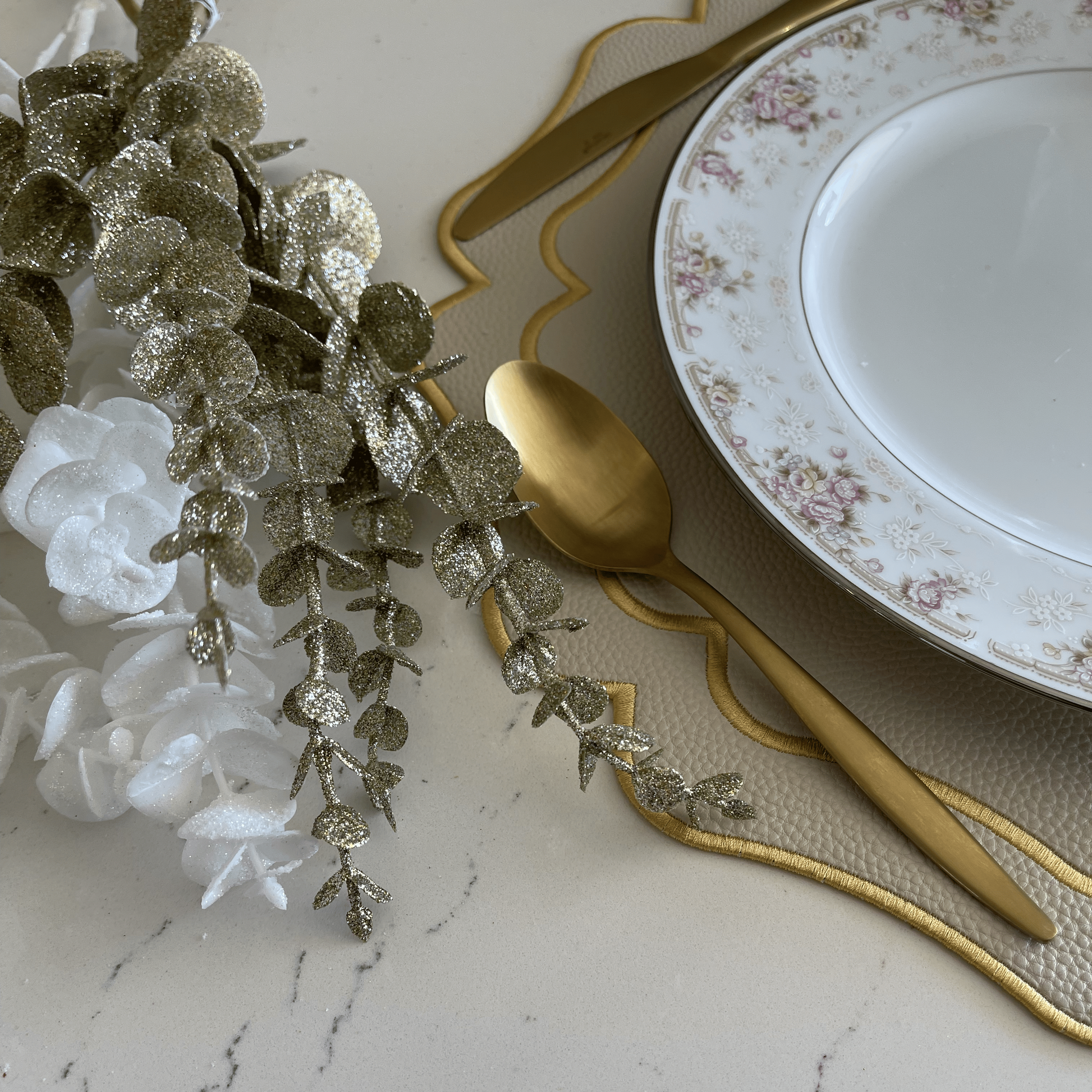 Decorative table setting with a white plate, gold cutlery, and greenery on a marble surface.