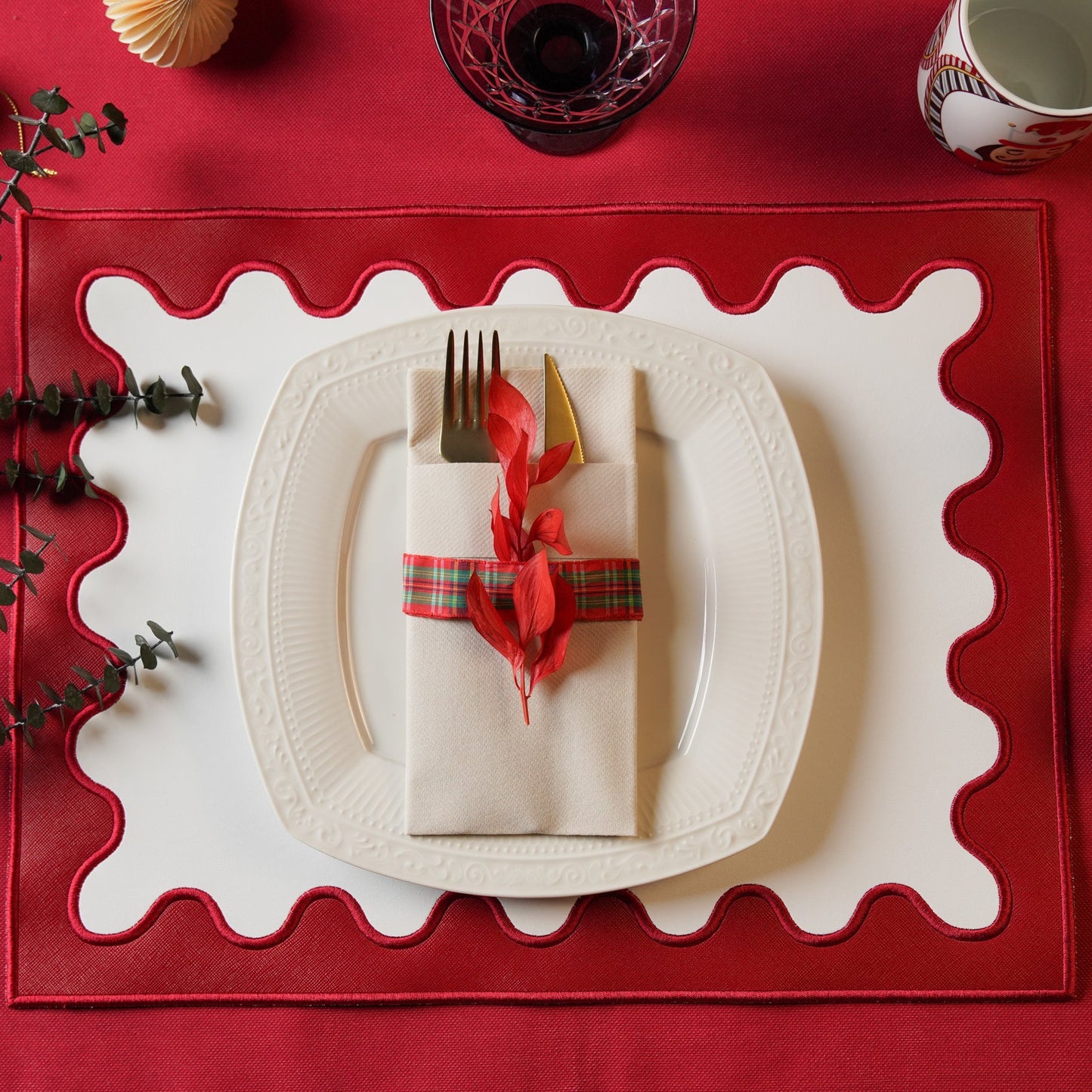 Dining table setting with a red placemat, white plate, and folded napkin with a red ribbon.