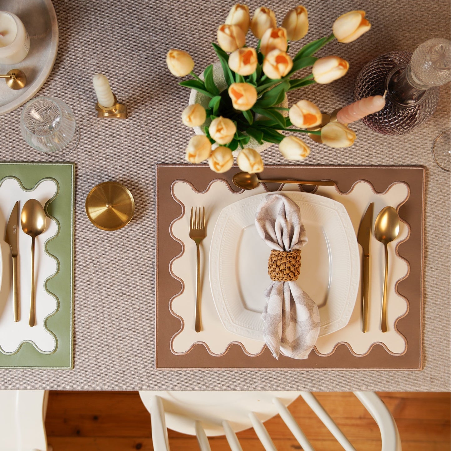 Dining table setting with gold cutlery, white plates, and a vase of yellow tulips on a beige tablecloth.