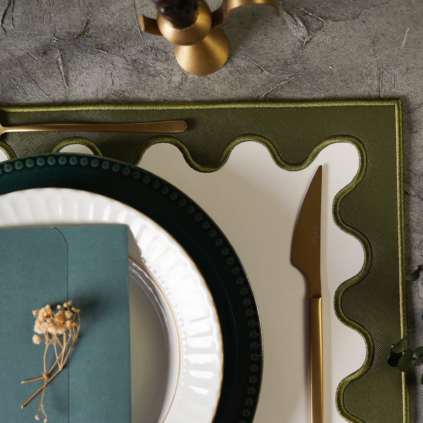 Elegant table setting with green charger, white plate, gold knife, and teal napkin on a gray surface.
