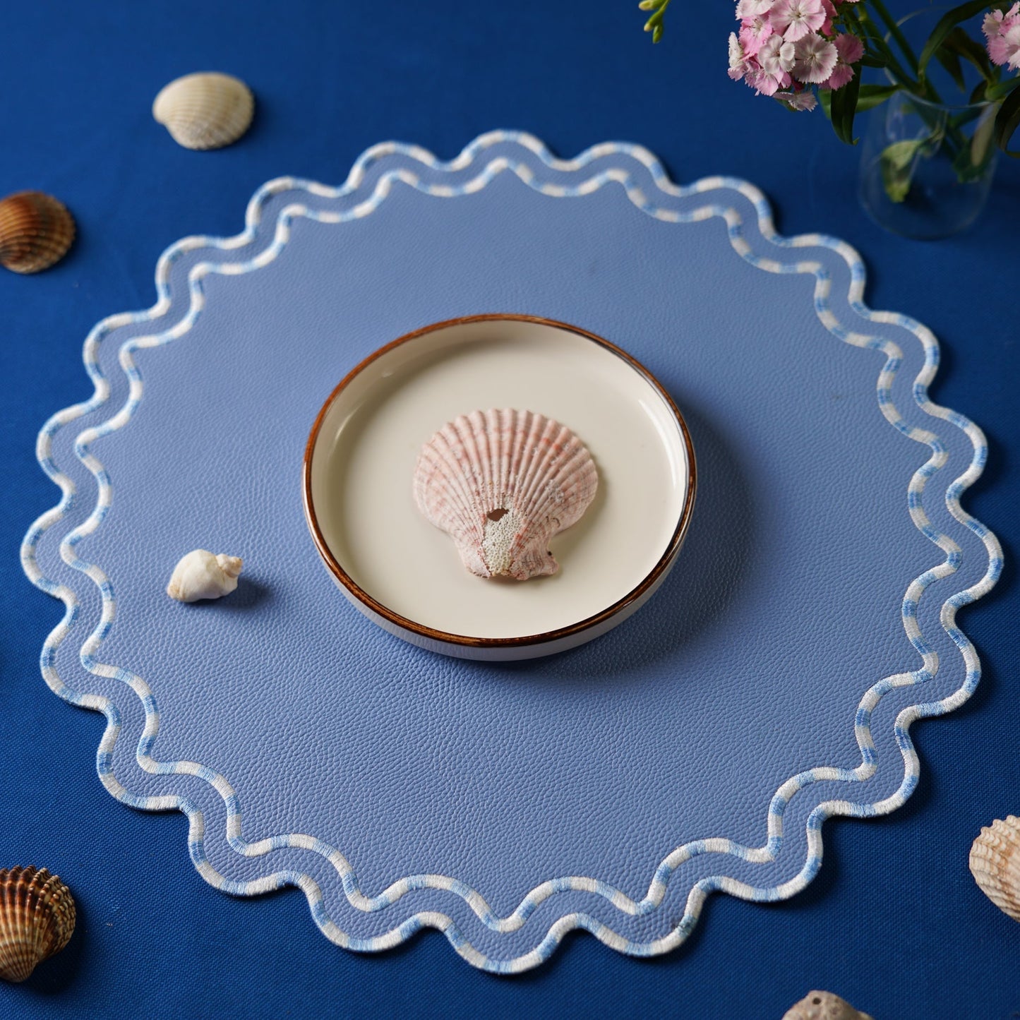 Decorative plate with a shell on a blue placemat, surrounded by seashells and flowers.