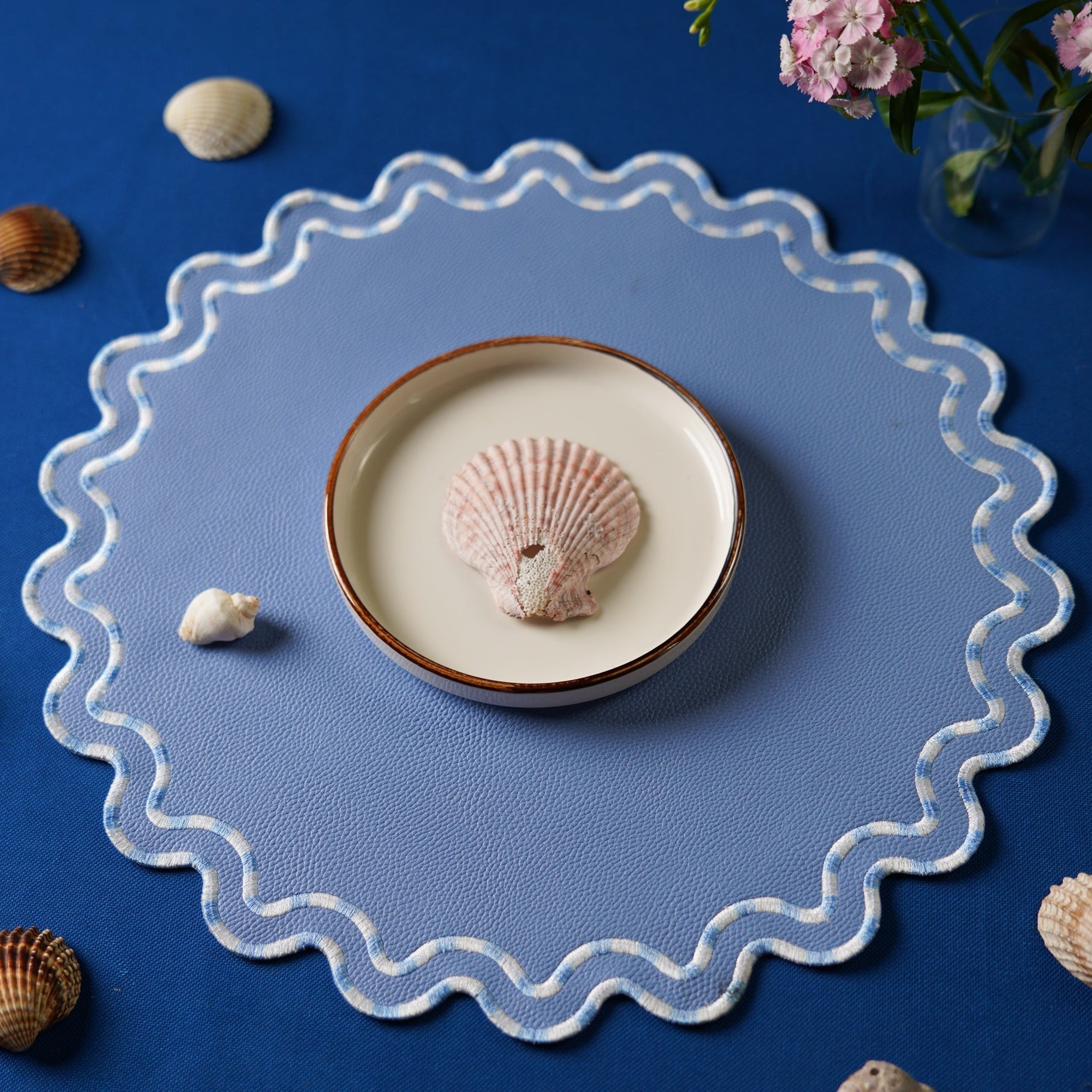 Decorative plate with a shell on a blue placemat, surrounded by seashells and flowers.